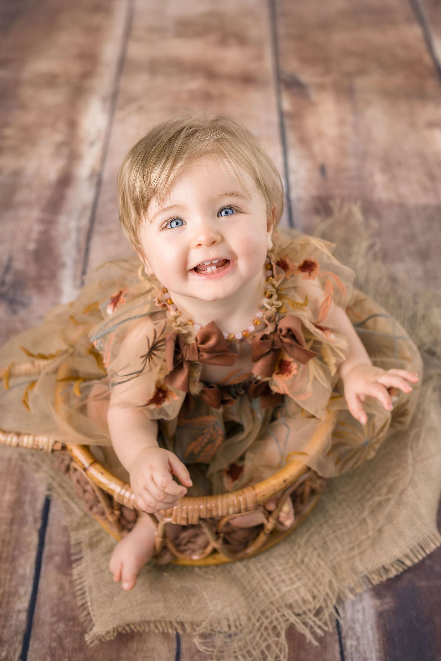 Happy blue-eyed baby girl in brown floral dress sitting in wicker basket on wooden floor.