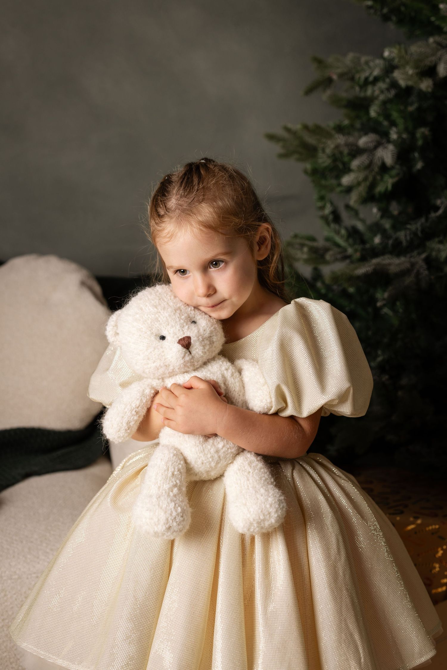 Young girl in cream dress hugging white teddy bear near Christmas tree.