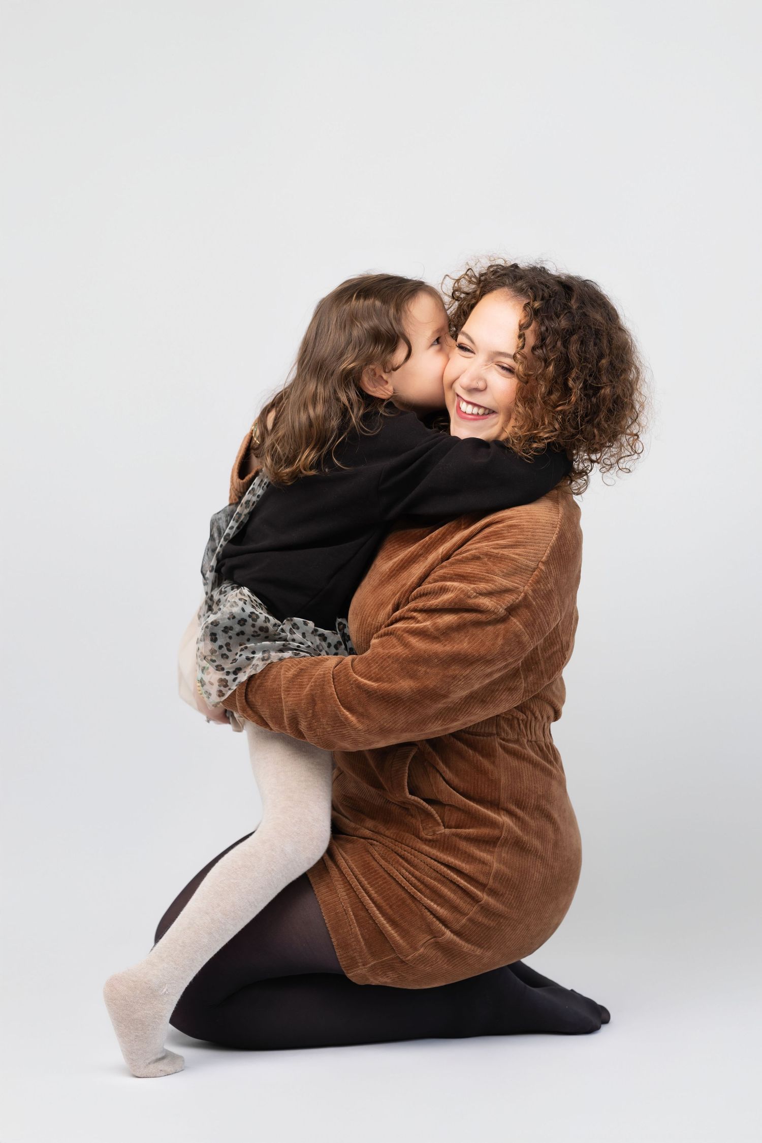 Joyful mother with curly hair laughing as young daughter gives her a kiss on the cheek.