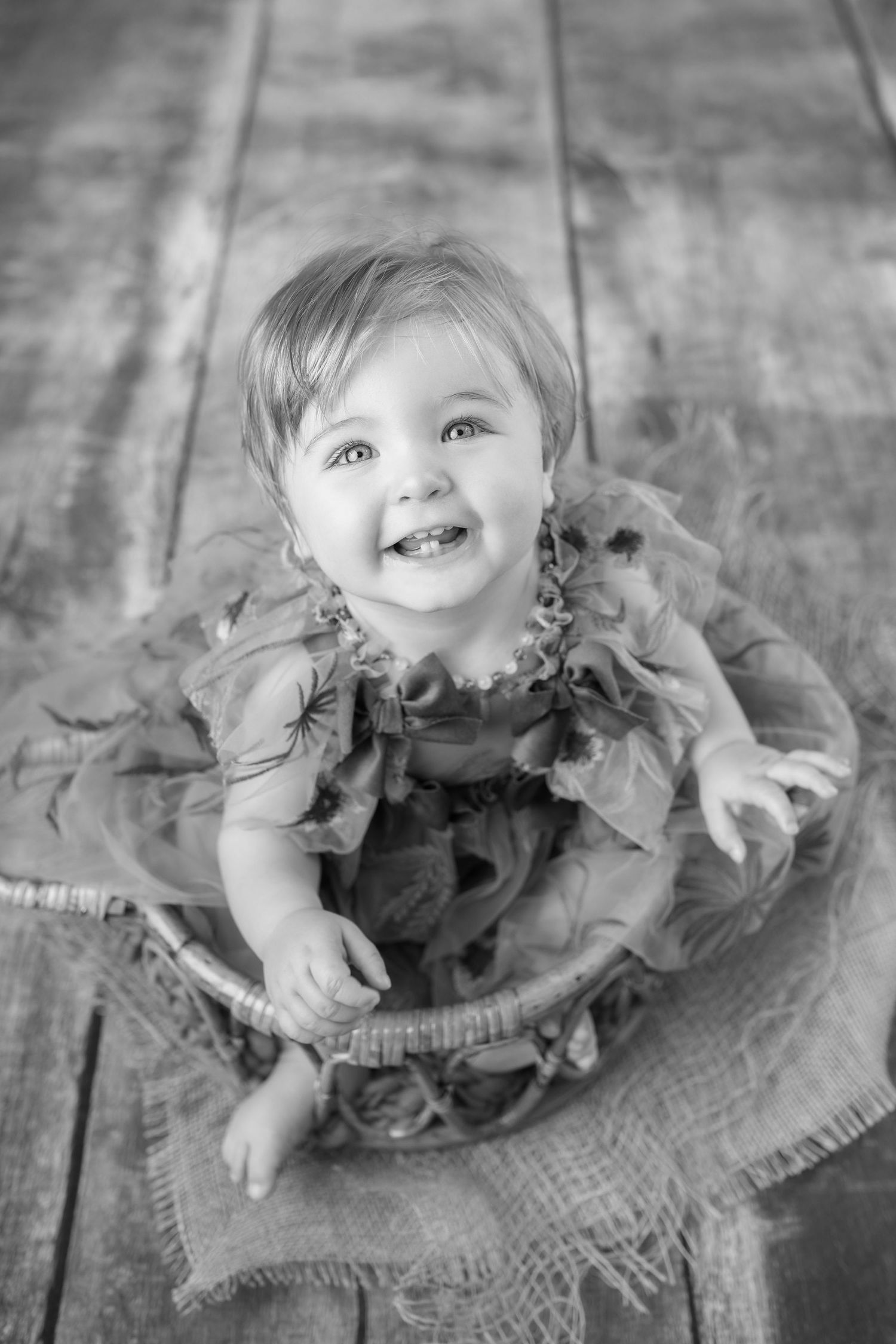 Black and white portrait of a happy baby girl sitting in a wicker basket wearing a ruffled dress.