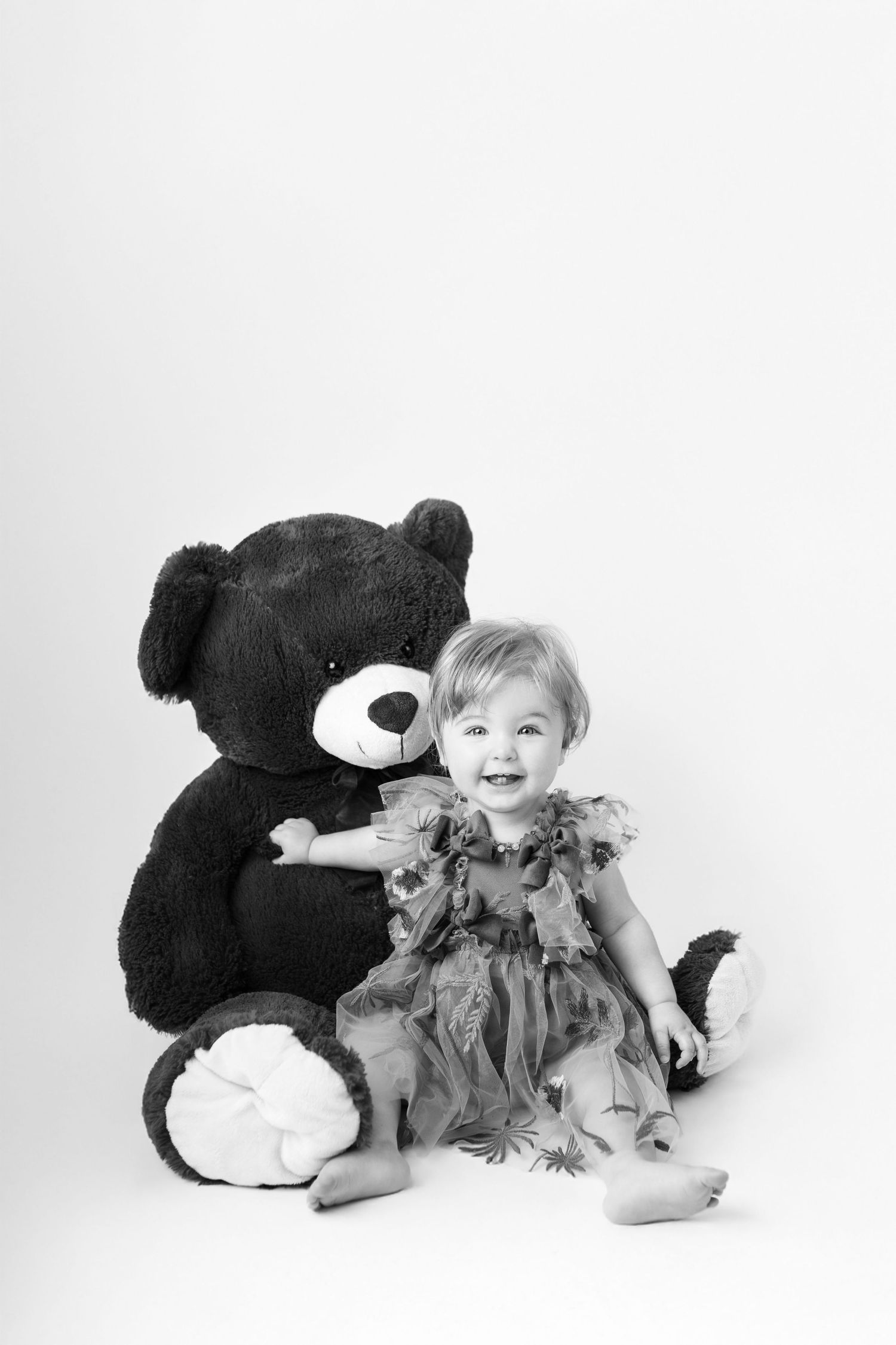 Laughing baby girl in a floral dress sits beside a large teddy bear in a black and white portrait.