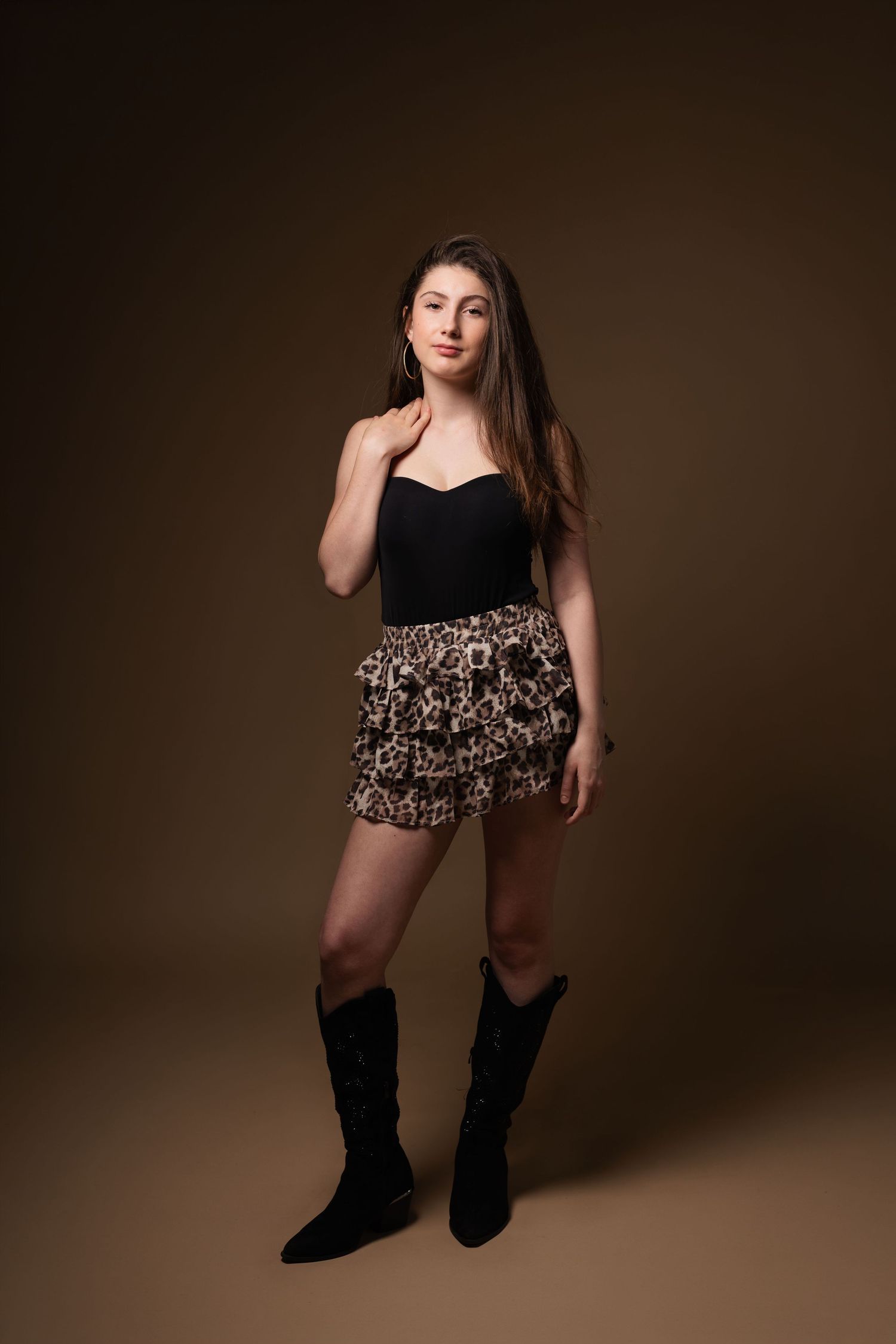 Young woman in black bustier, leopard print mini skirt, and black cowboy boots posing against brown studio backdrop.