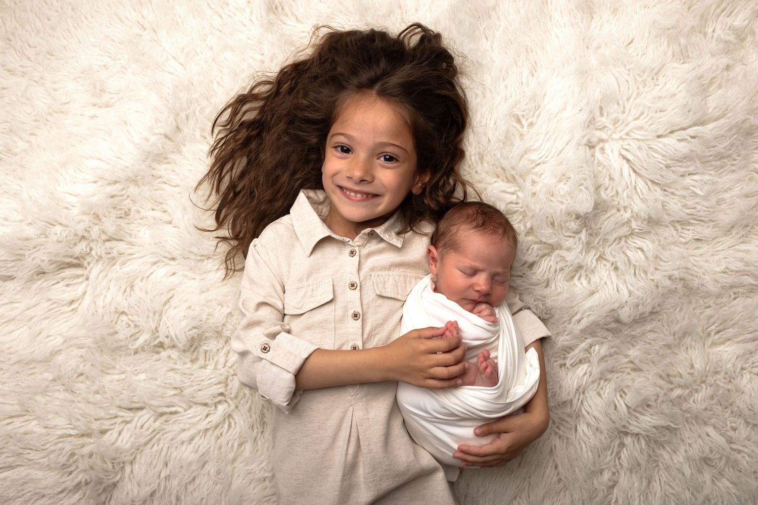 Young girl with curly hair smiles while holding a swaddled newborn baby on a fluffy white fur background.