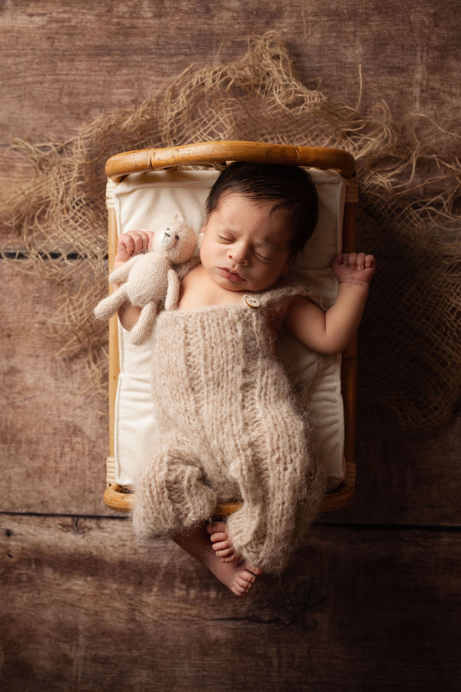 Newborn baby sleeping in tiny wooden bed wearing knit romper, holding small stuffed animal toy on rustic wood background.