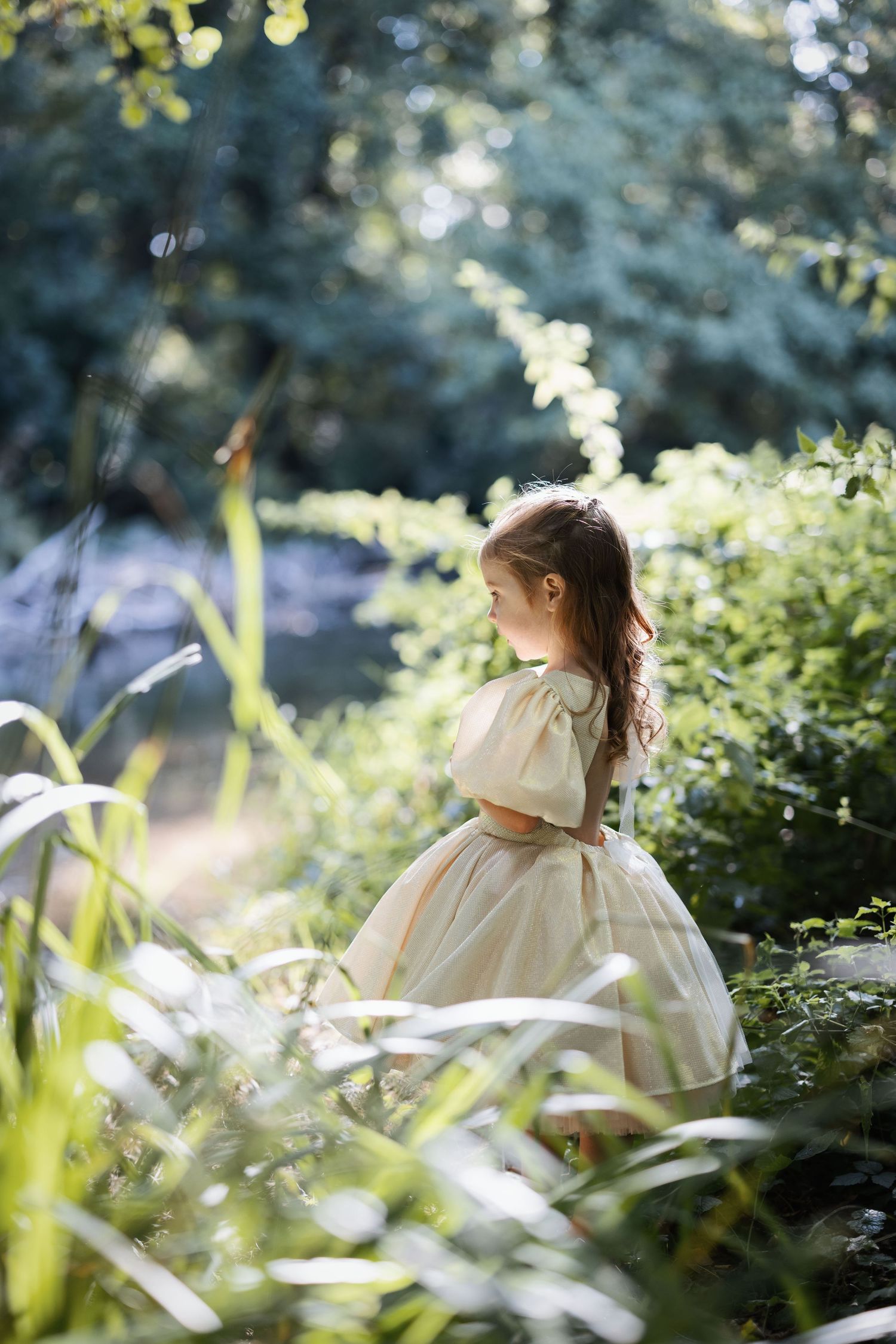 A young girl in a cream ball gown dress stands among lush green garden foliage in soft natural sunlight.