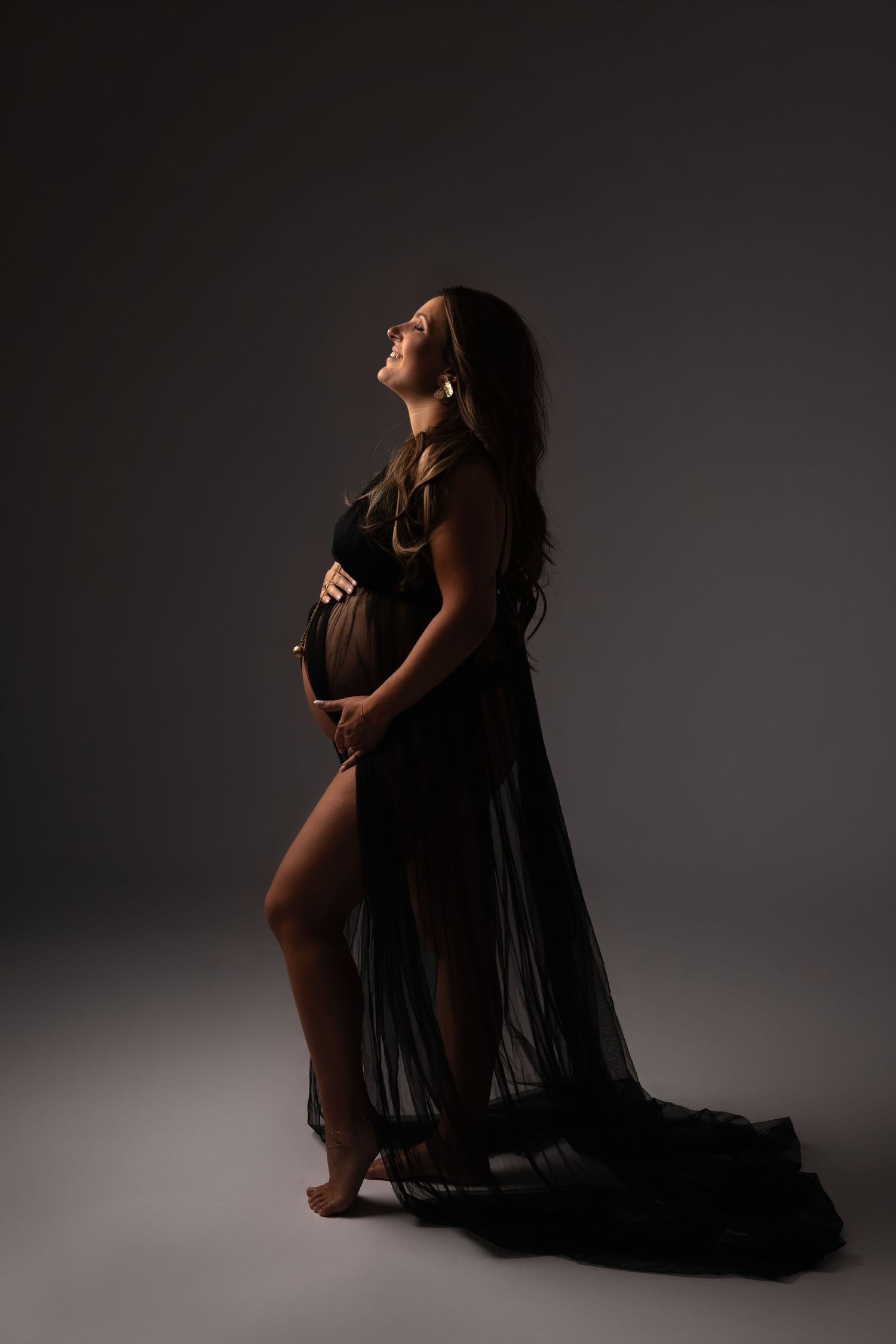 Pregnant woman in sheer black gown cradling baby bump, elegant maternity studio portrait with dramatic lighting.