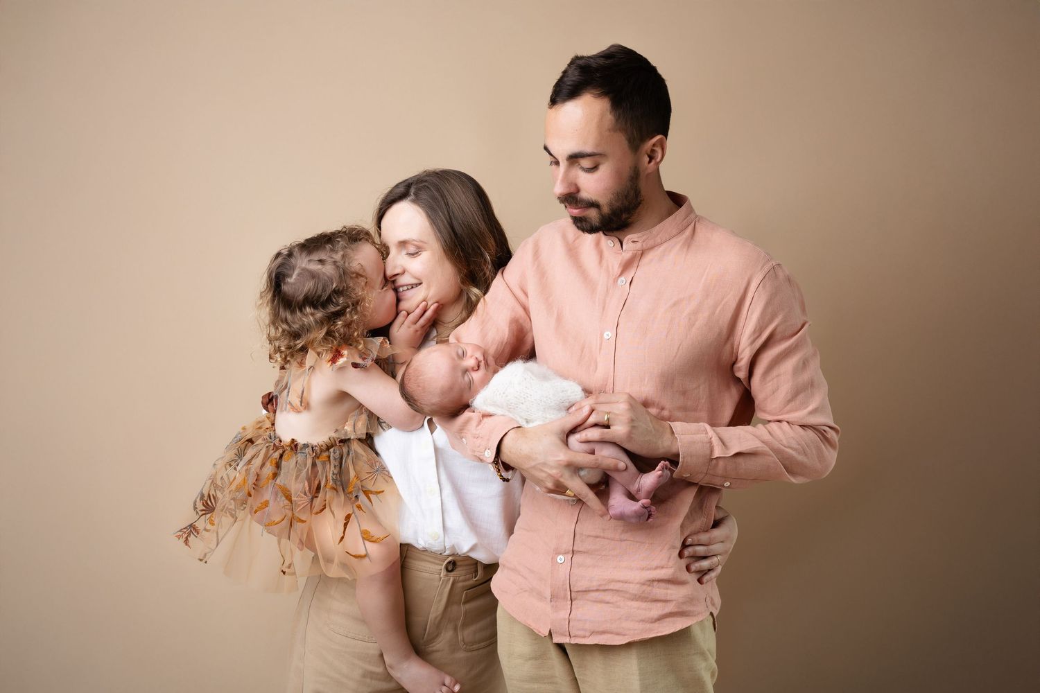 Young family with newborn baby and toddler daughter posing together in neutral tones studio portrait.