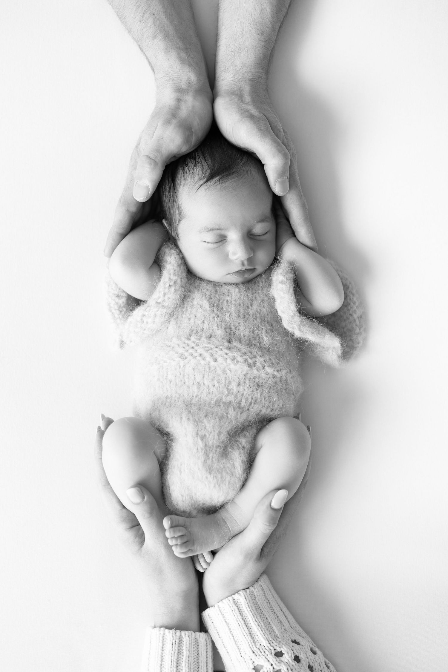 Parents hands gently cradling sleeping newborn baby dressed in knit outfit, black and white.
