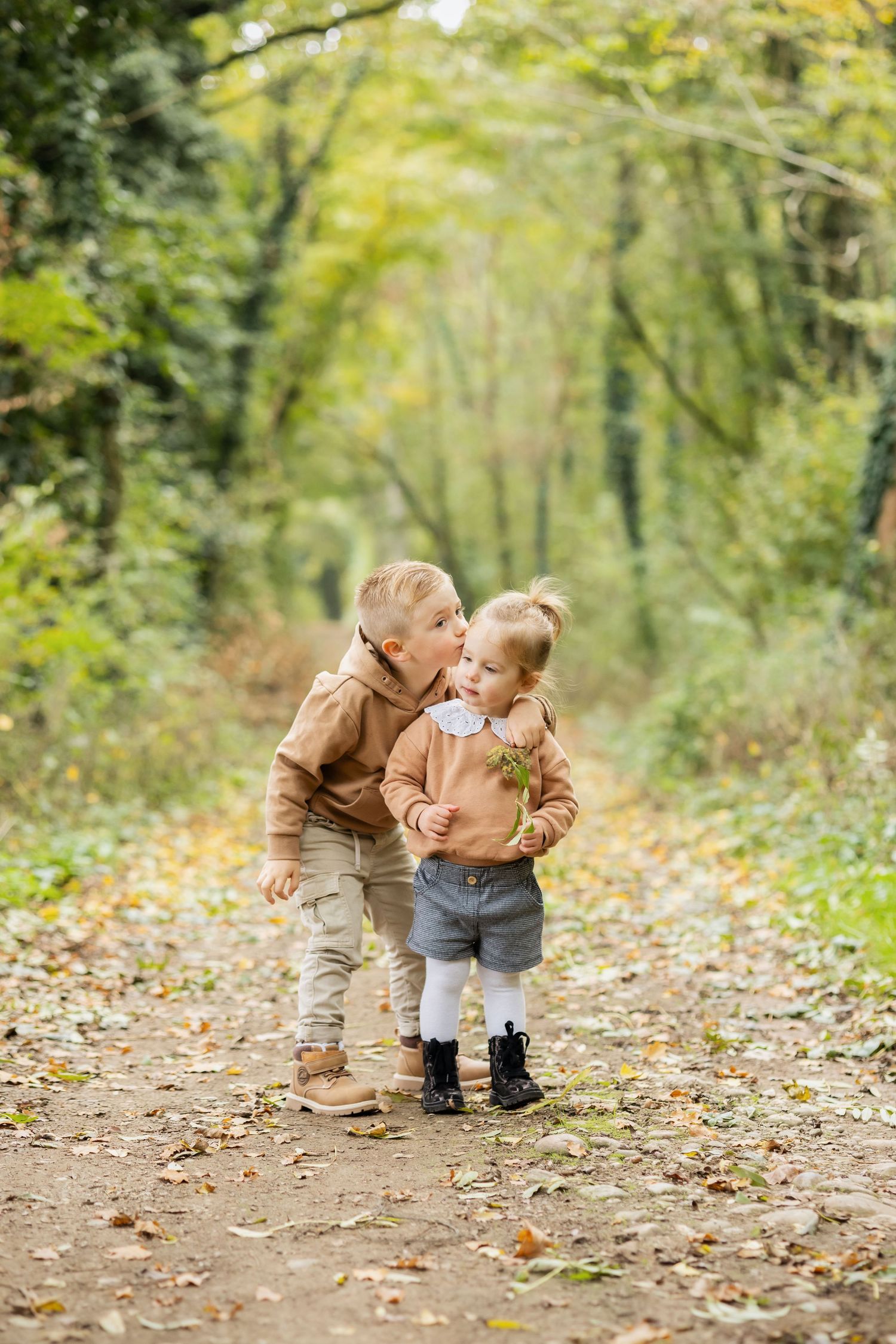 Two young children share a sweet moment on a leaf-covered forest path in autumn, dressed in matching beige outfits.