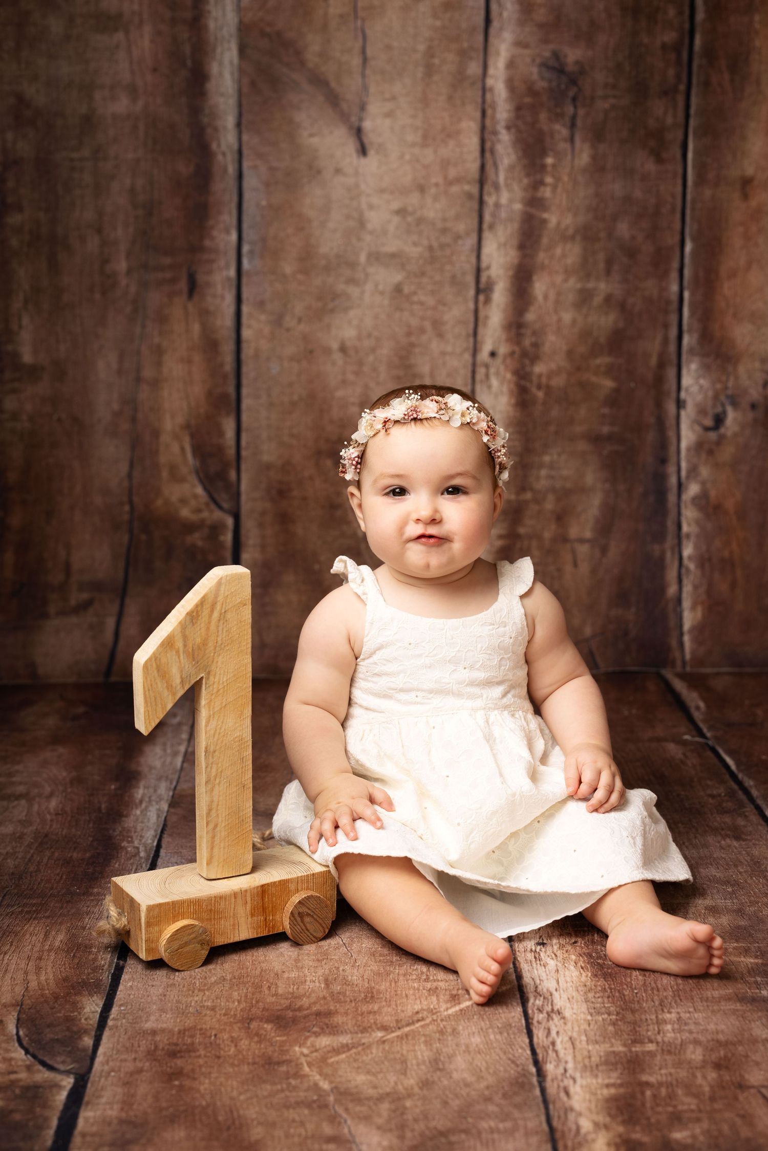 Baby girl in white dress with floral headband sits beside wooden number one toy on rustic wood floor.