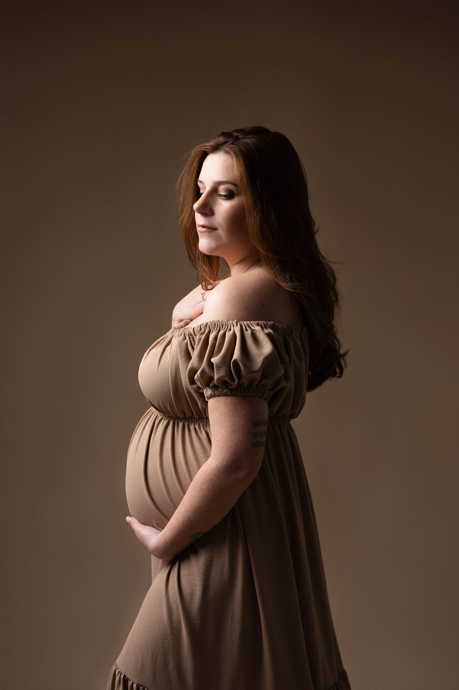Pregnant woman in off-shoulder beige dress holding her belly in a warm-toned studio. photographe grossesse chavanoz lyon