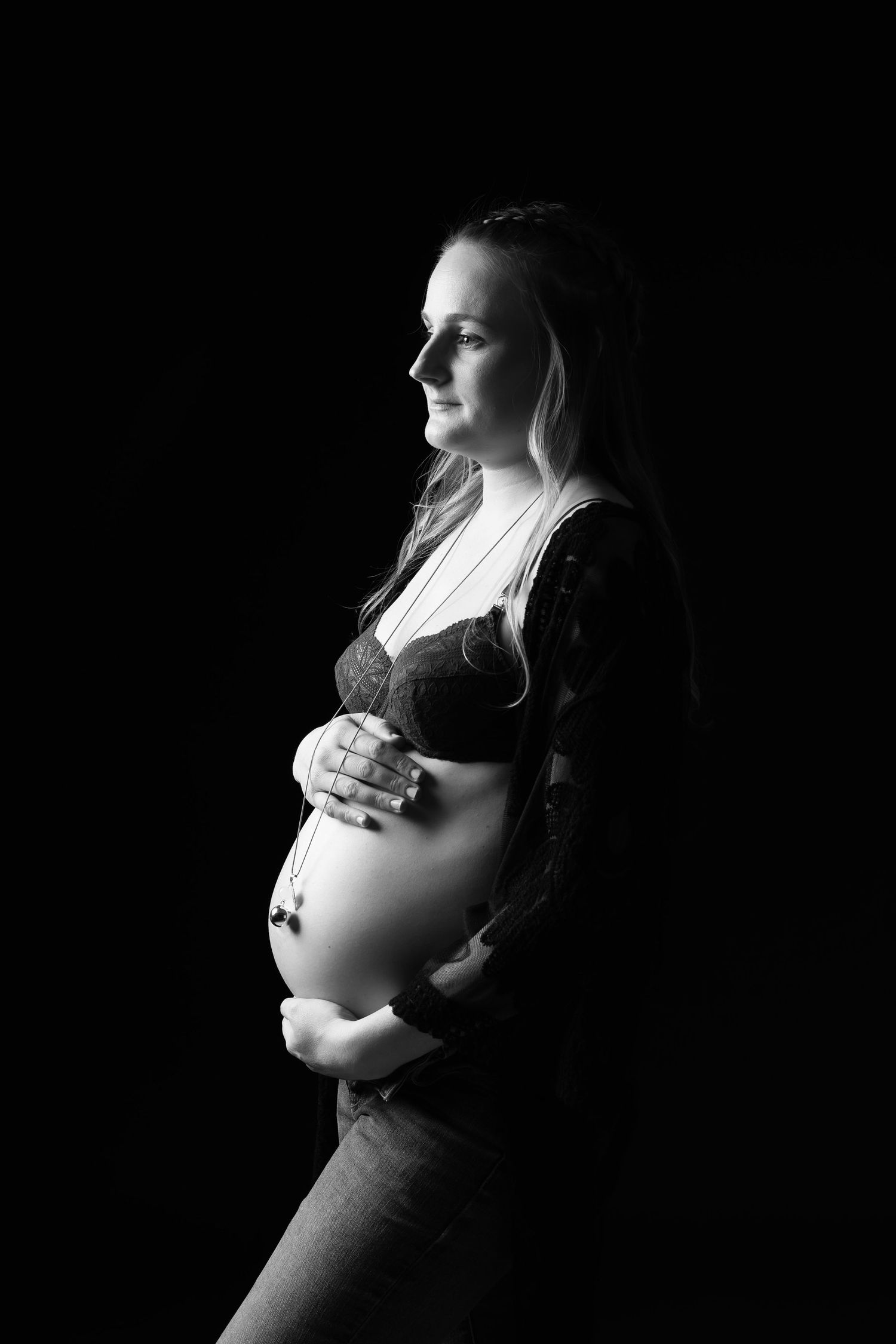 Black and white profile portrait of pregnant woman cradling bare belly against dark background. photographe grossesse chavanoz lyon