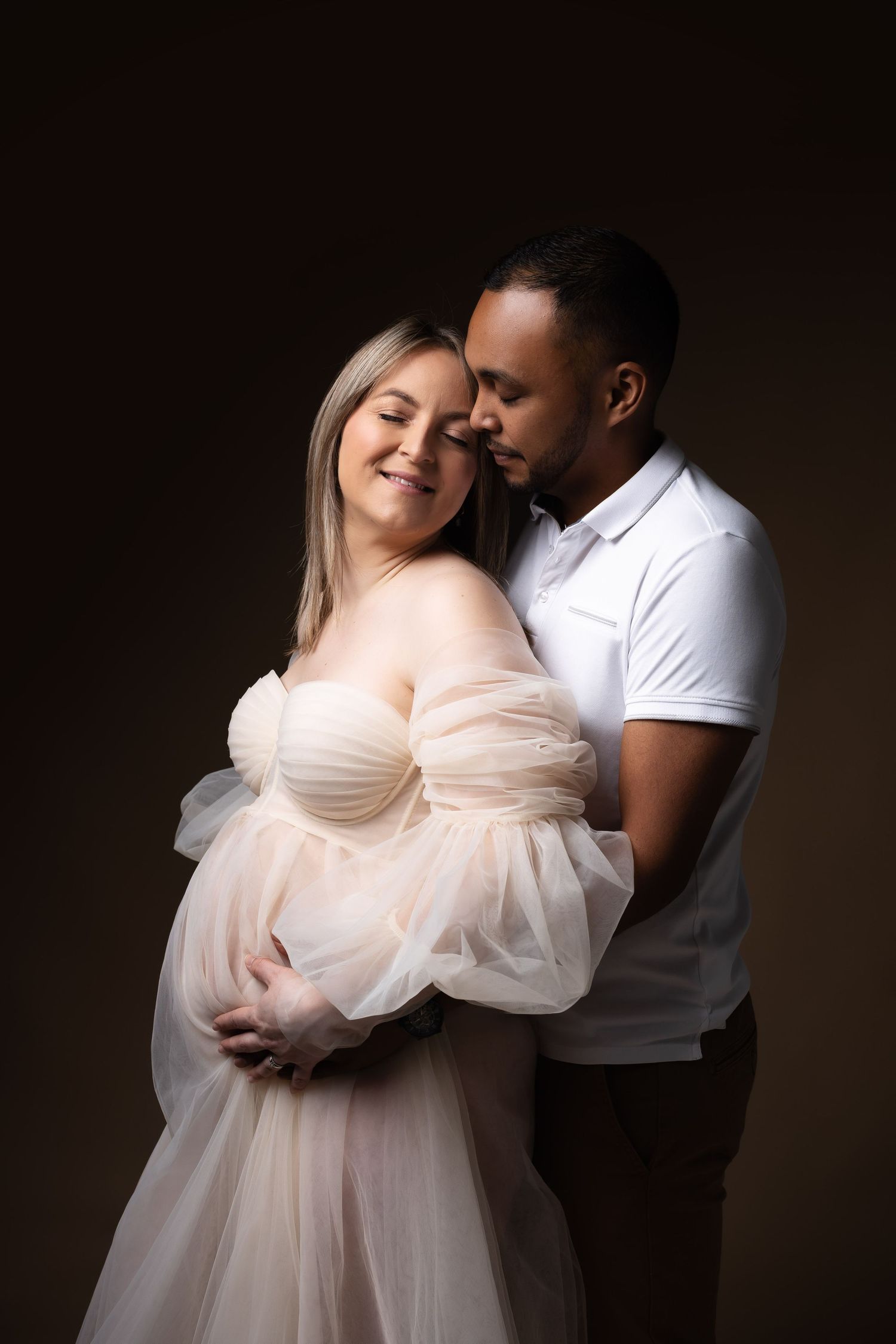 Smiling pregnant woman in sheer tulle dress embraced from behind by partner on dark studio background.