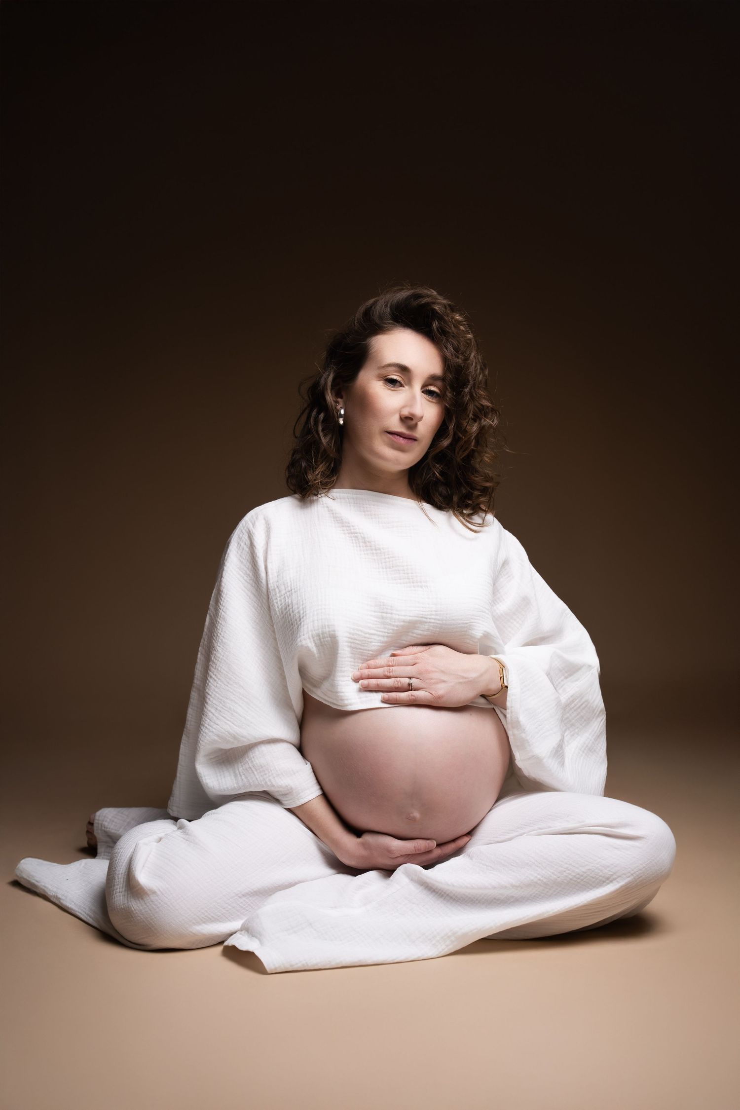 Pregnant woman in white top sits cross-legged on studio floor, lovingly holding her exposed baby bump.