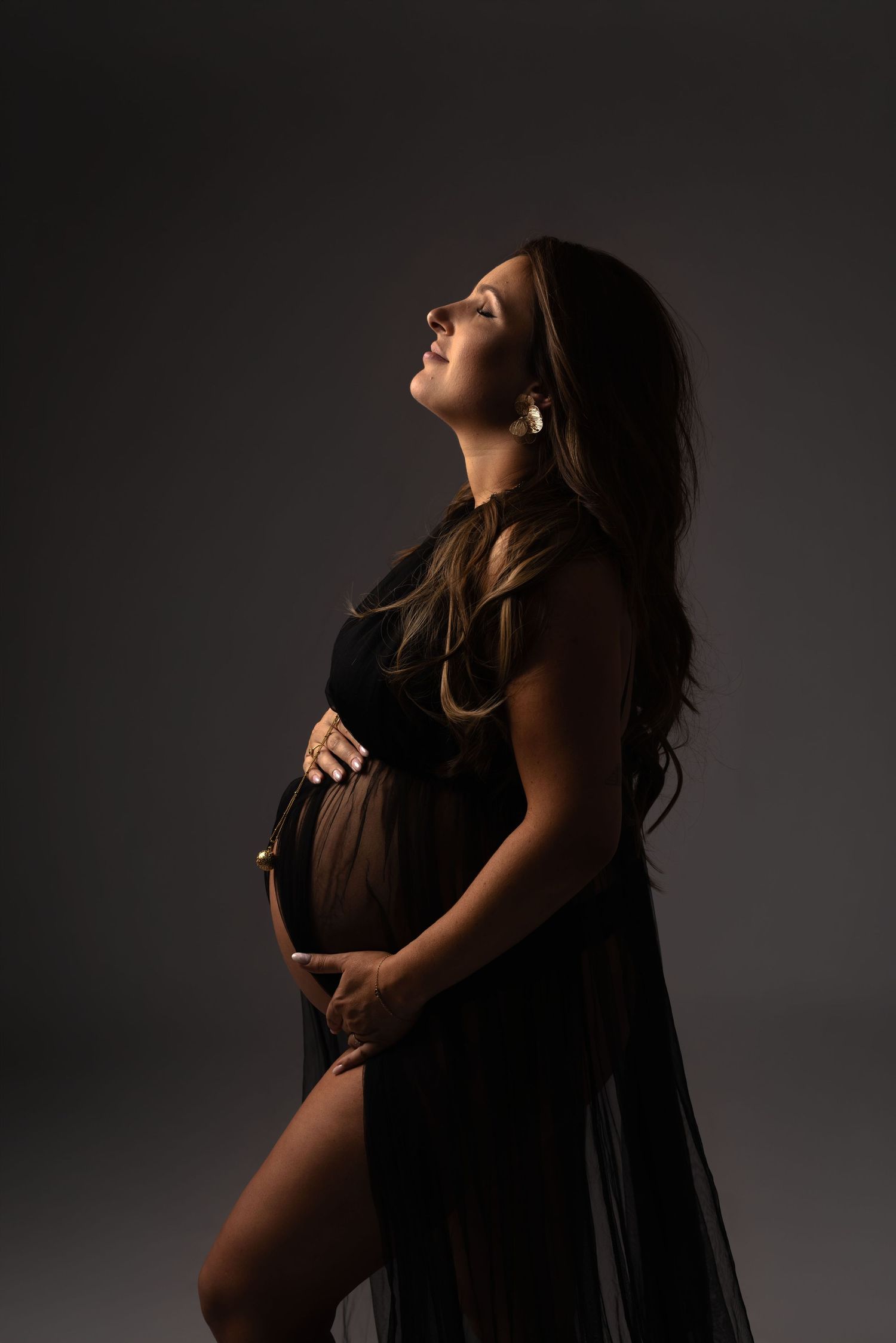 Pregnant woman in black sheer gown cradling her baby bump, looking up gracefully against a dark studio background.