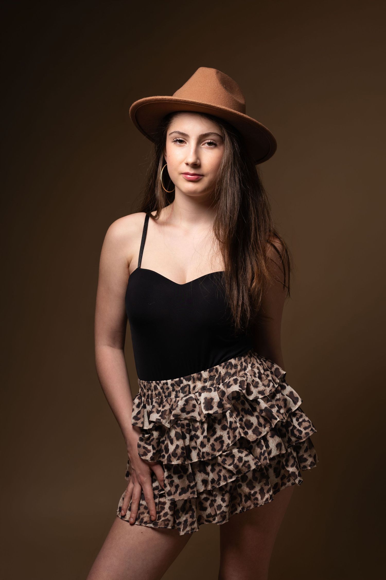 Young woman in brown fedora hat, black cami top, and leopard print ruffled mini skirt posing against brown background.