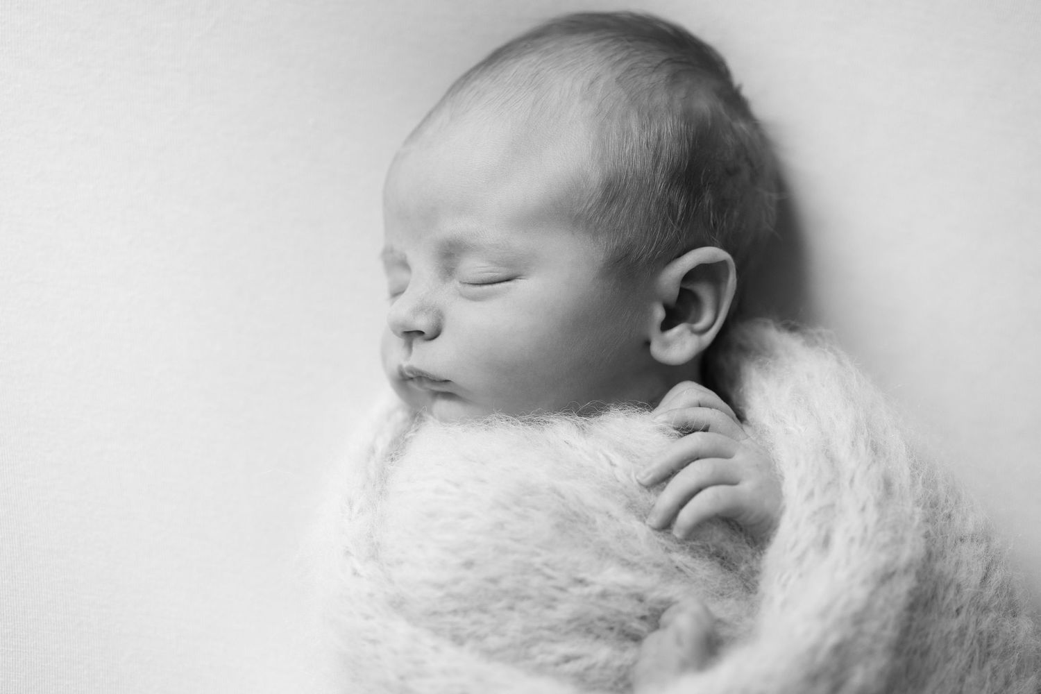Peaceful sleeping newborn baby wrapped in soft fluffy white knit blanket, black and white portrait photography.