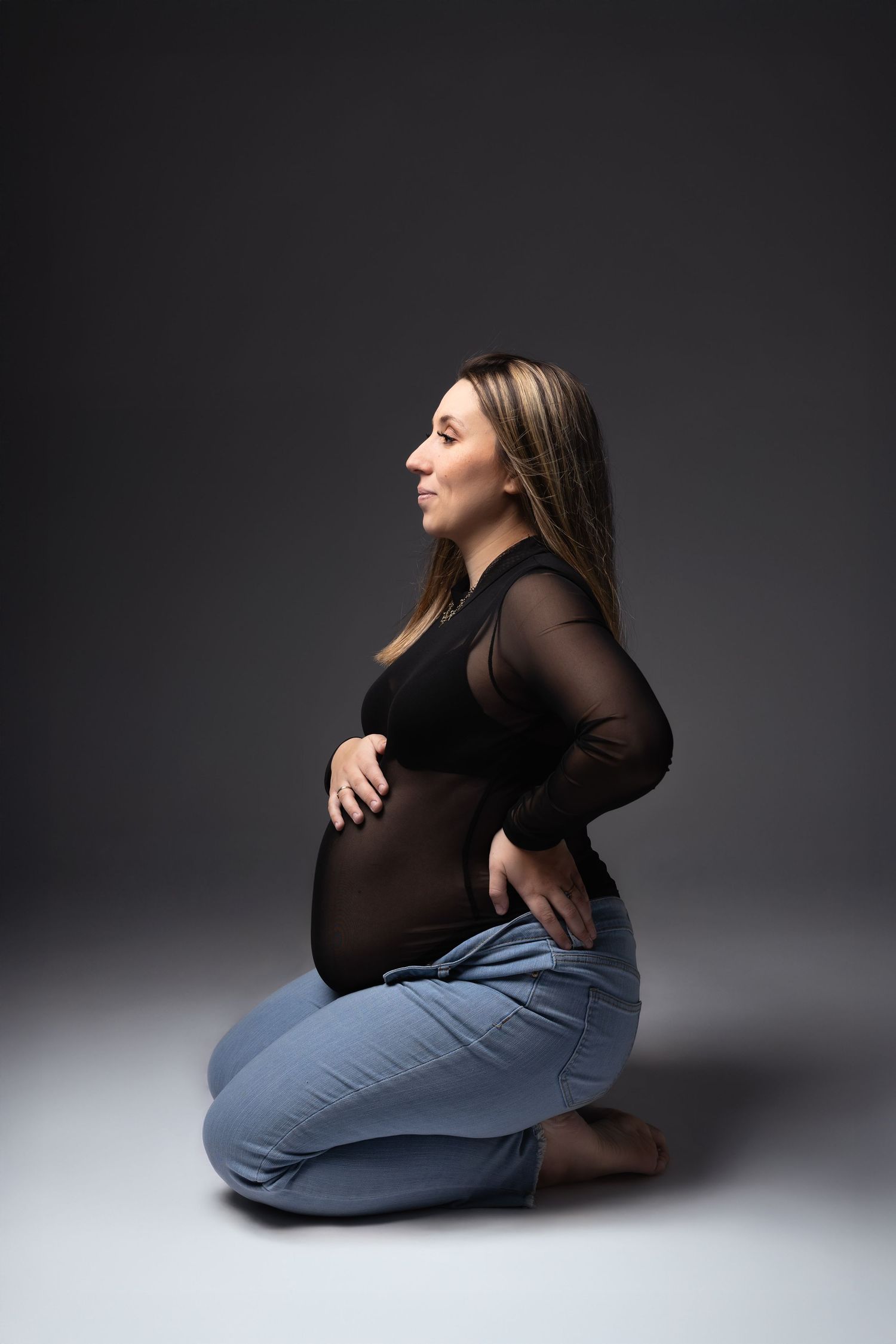 Pregnant woman in black sheer top and jeans kneeling gracefully in studio. photographe grossesse chavanoz lyon