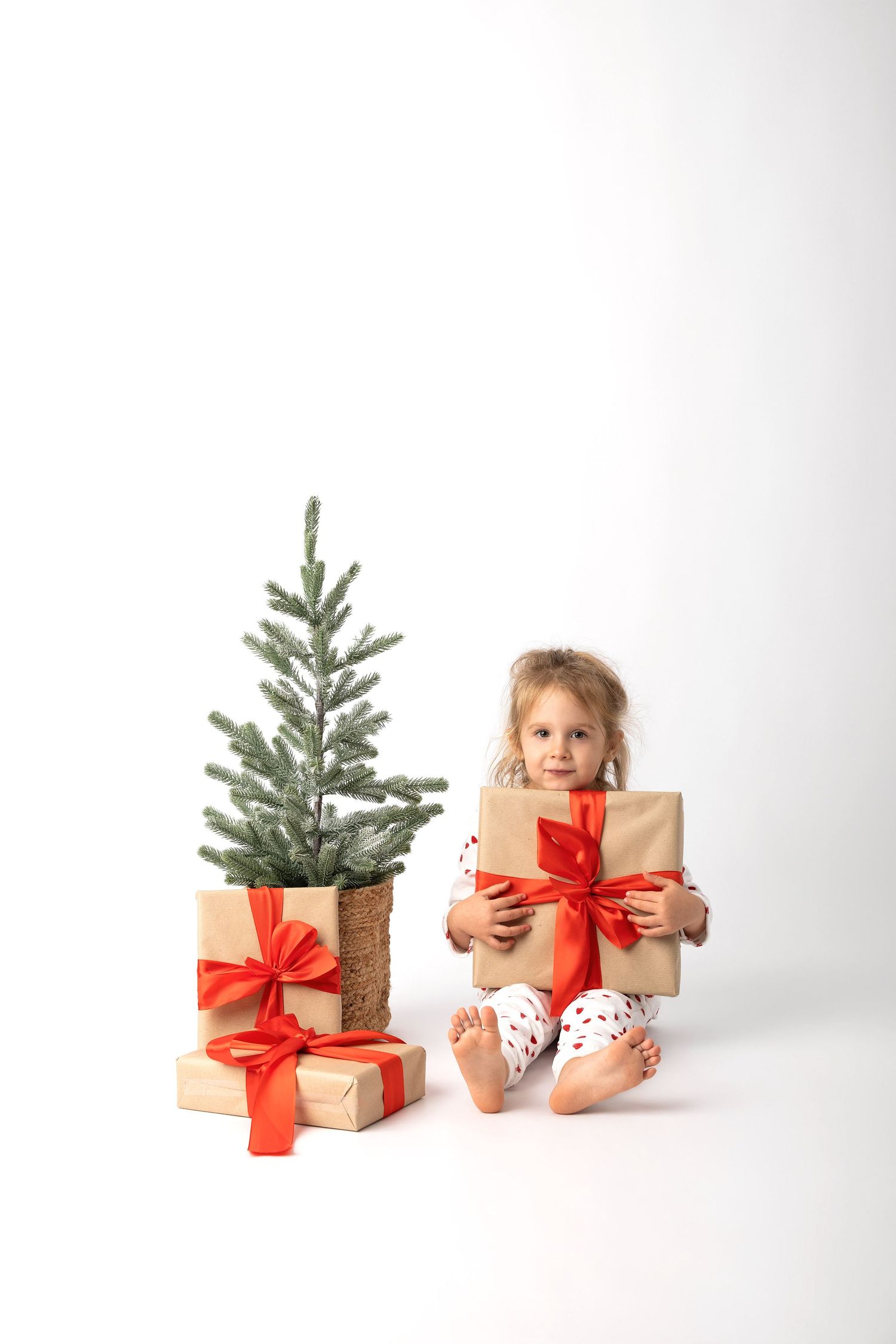 Young girl in pajamas sitting on floor holding Christmas gift boxes with red ribbons next to small decorated tree.