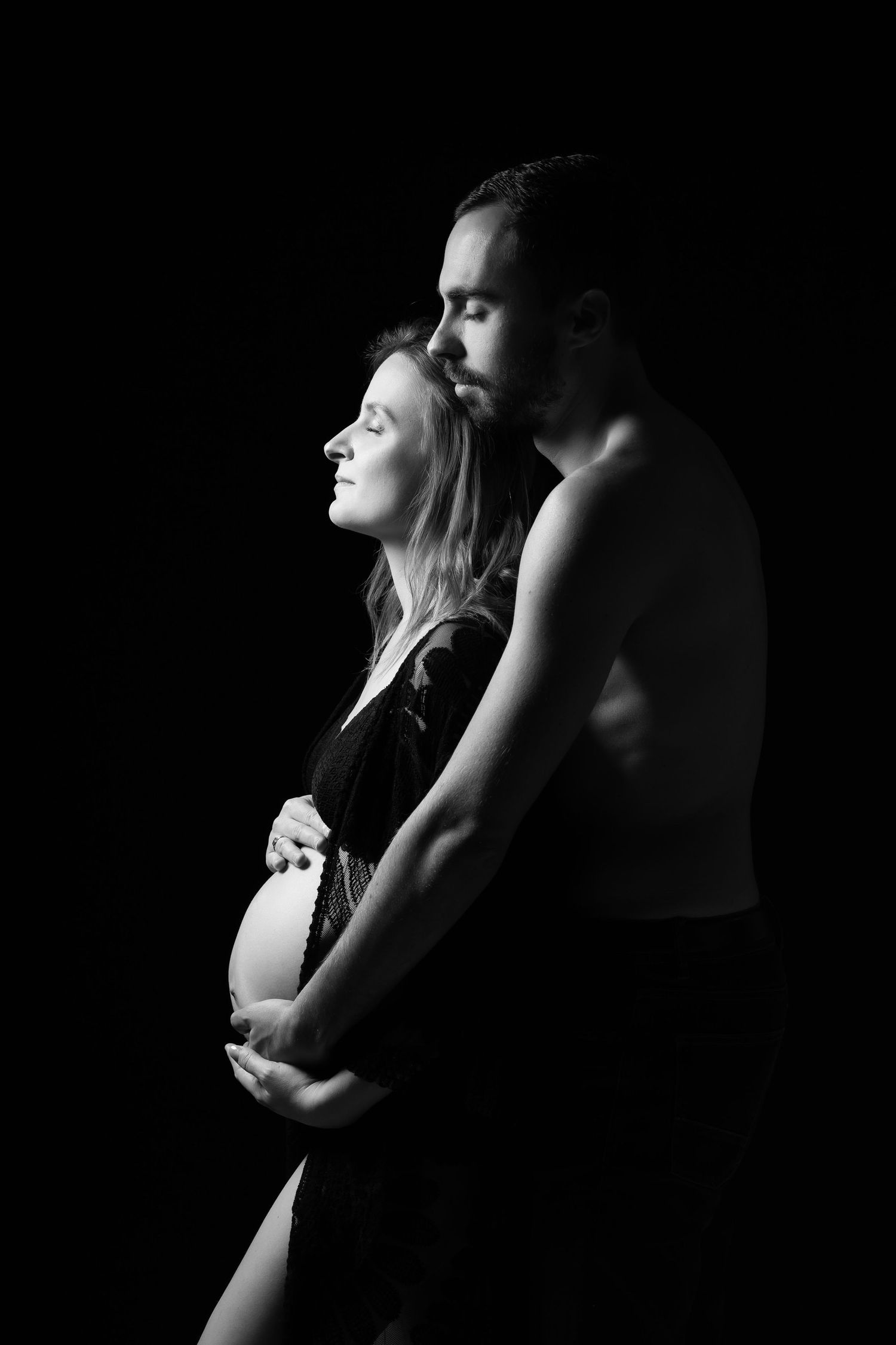 Black and white silhouette of shirtless man embracing pregnant woman against dark background.