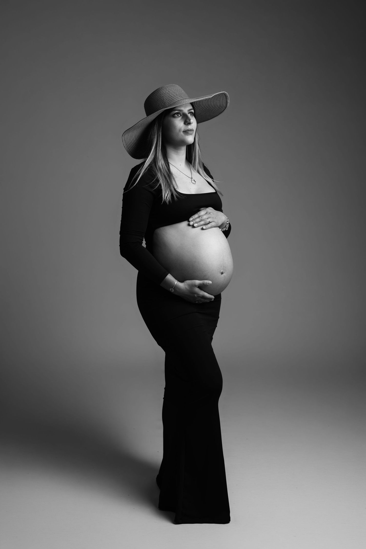 Black and white maternity photo of pregnant woman in black outfit and wide-brim hat cradling her baby bump in studio.