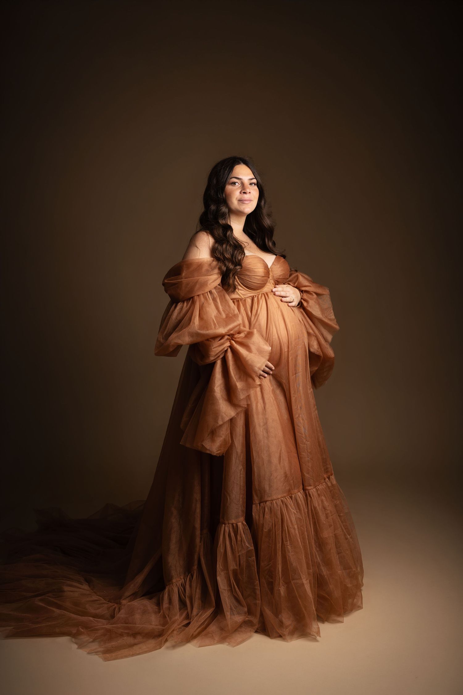 Pregnant woman in elegant brown tulle maternity gown posing gracefully in a professional studio portrait.