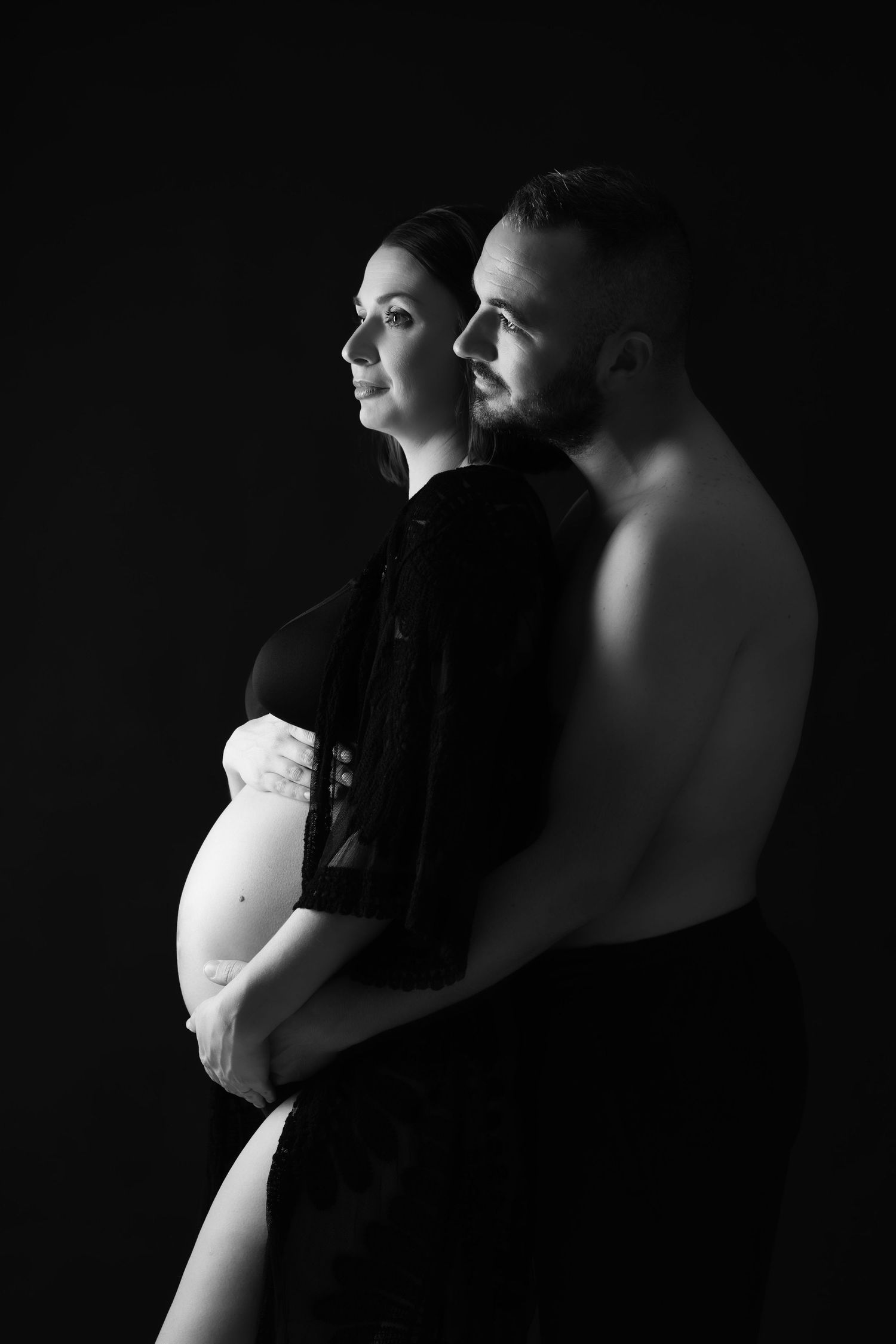 Black and white artistic portrait of pregnant couple embracing in profile on dark background.
