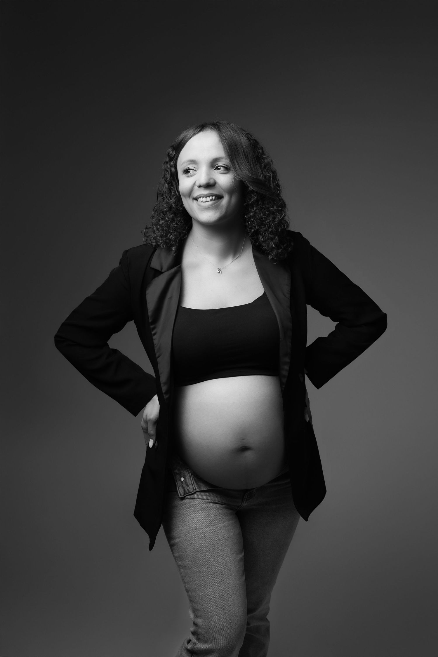 Black and white portrait of smiling curly-haired pregnant woman in blazer with hands on hips.