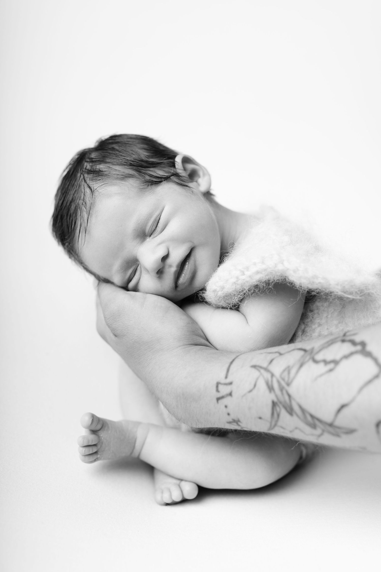 Black and white photo of a sleeping newborn baby cradled in tattooed arms, smiling peacefully.