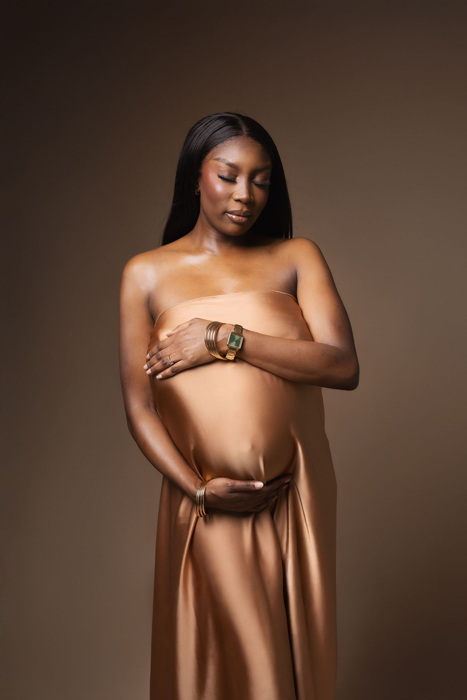 Pregnant Black woman wrapped in gold satin with bracelet in studio.