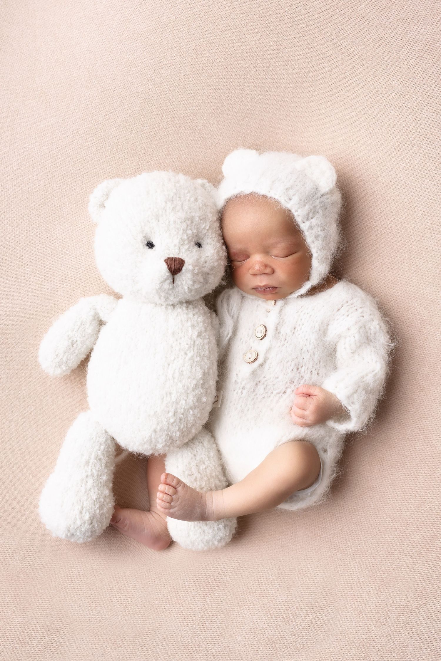 Sleeping newborn in white fuzzy bear outfit cuddling a white teddy bear.