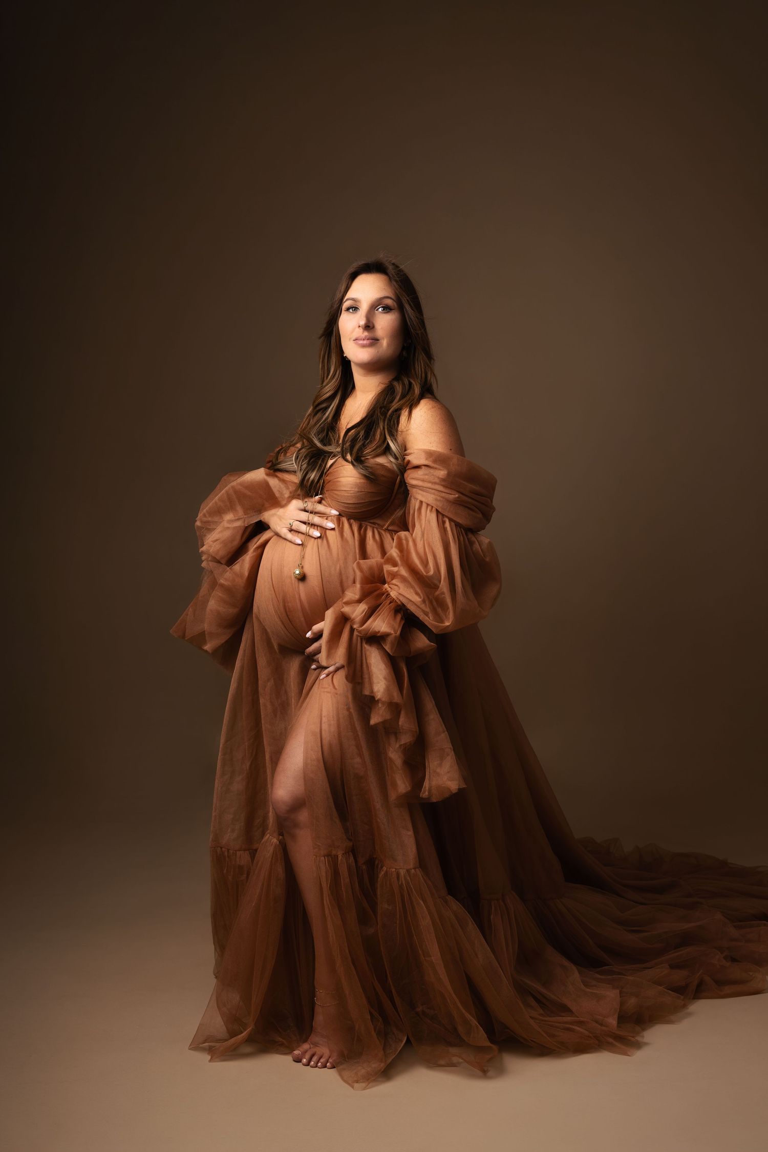 Pregnant woman in flowing rust tulle maternity gown cradling belly in professional studio portrait.