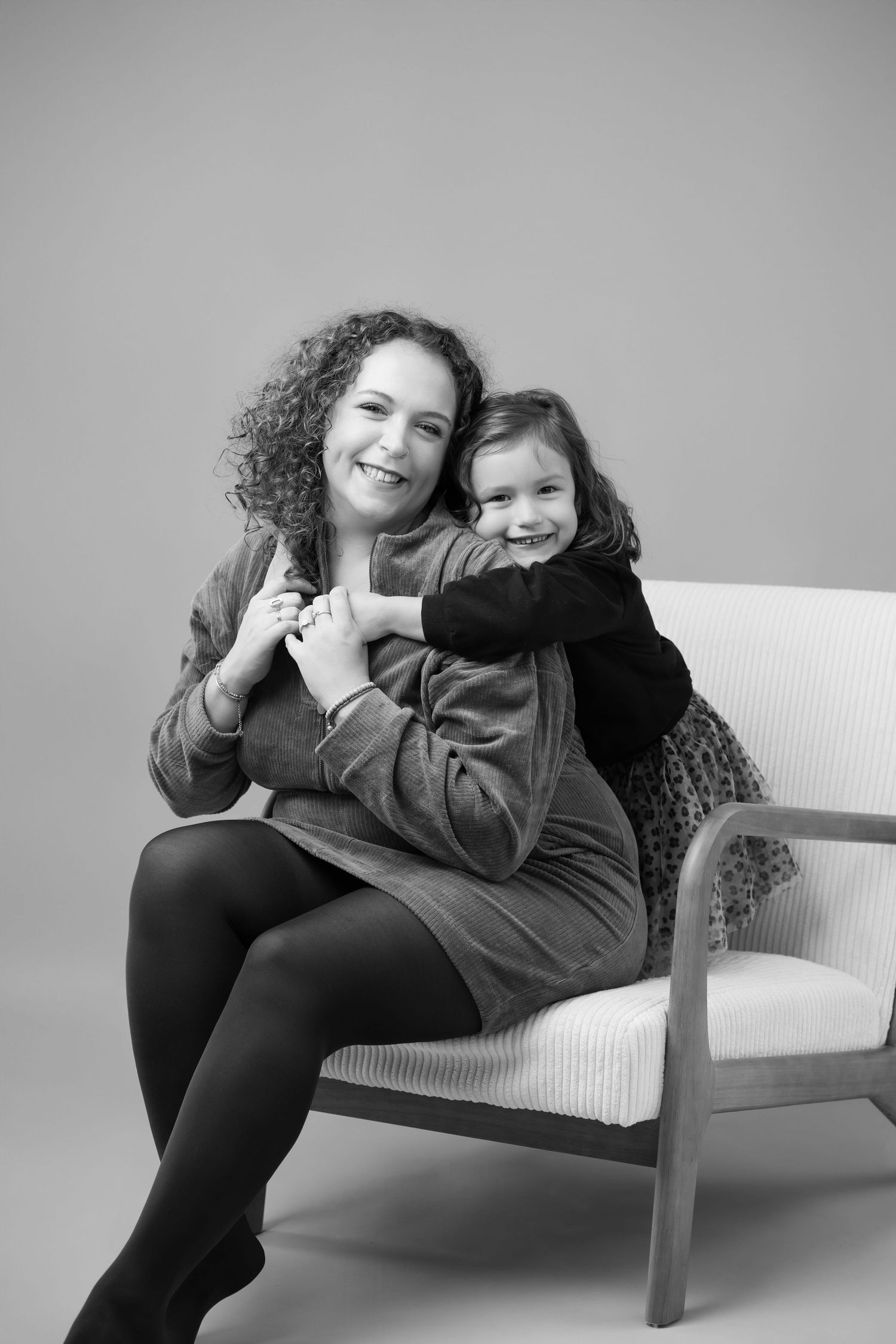 Black and white portrait of smiling mother seated in chair with daughter hugging her from behind.