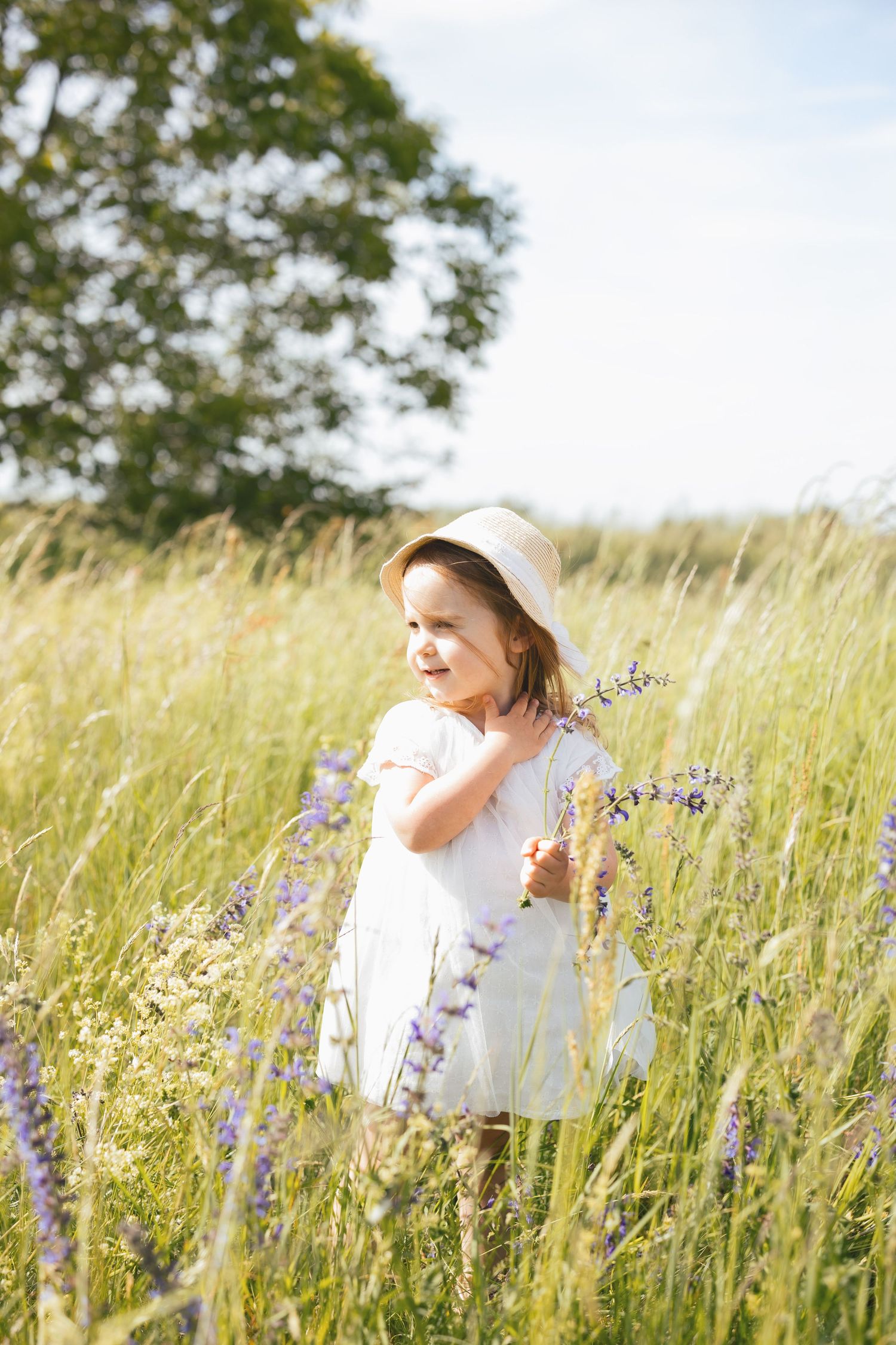 Young girl in white dress and straw hat holding lavender flowers in a sunny meadow field.