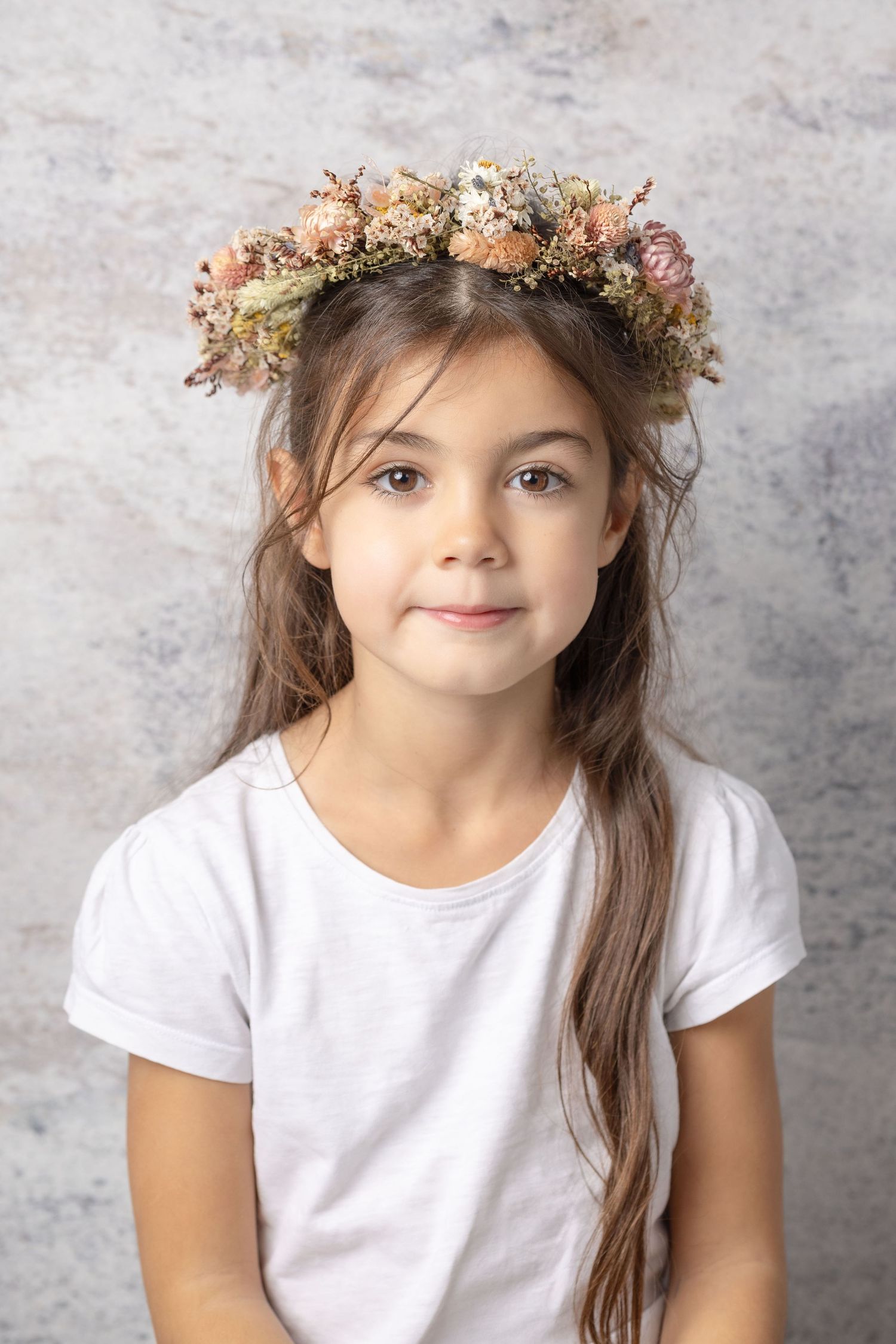 Young girl with long brown hair wearing a delicate dried flower crown and white t-shirt in studio portrait.