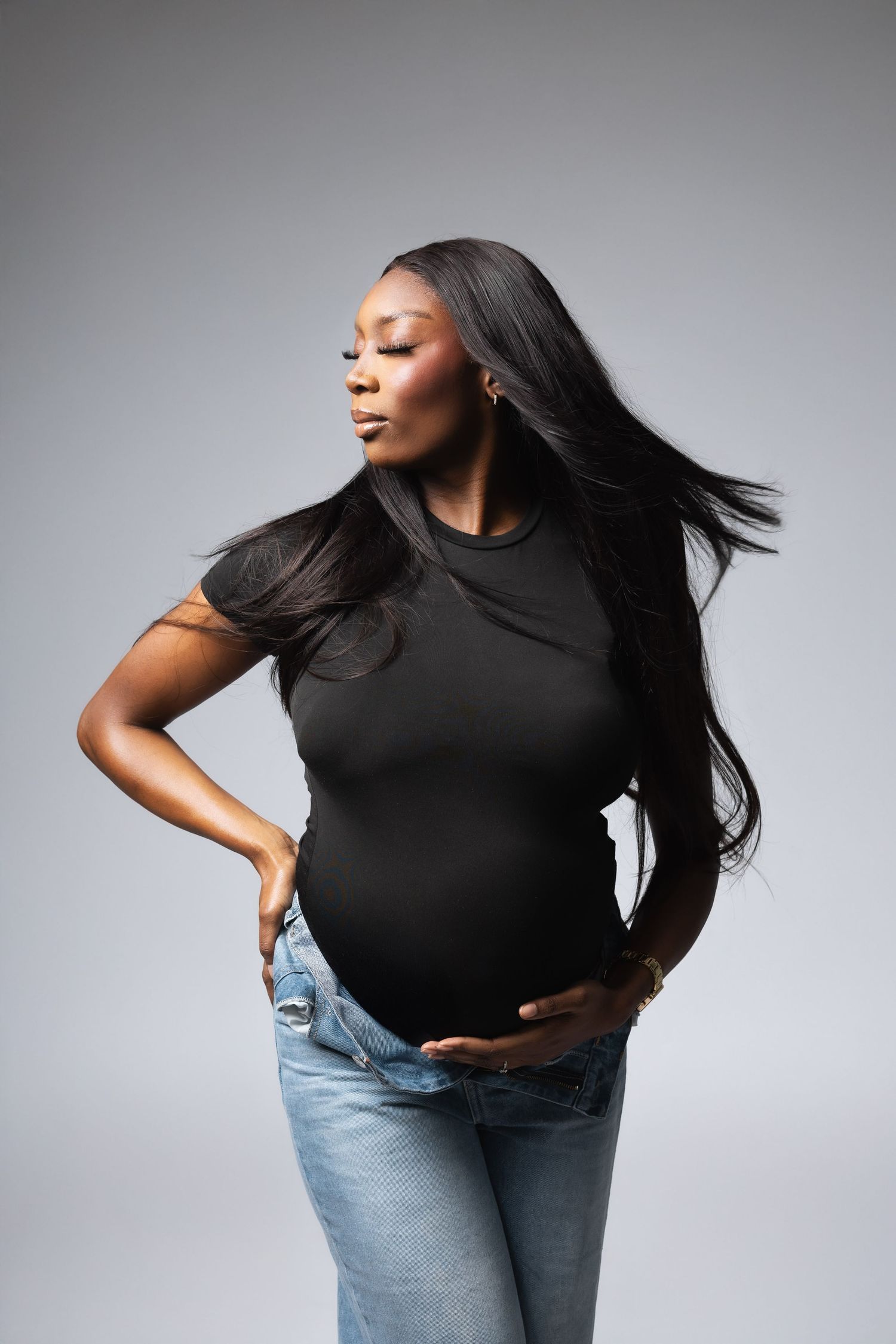 Pregnant Black woman in black tee and jeans with flowing hair in studio.