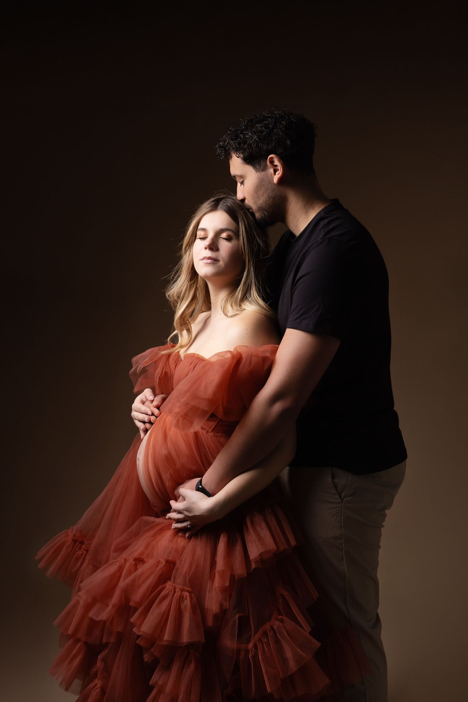 Couple sharing tender maternity moment, man embracing pregnant woman in red tulle dress against brown backdrop.