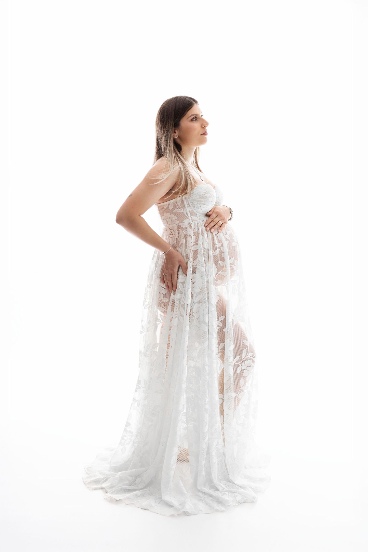 Pregnant woman in elegant white lace maternity gown posing gracefully against a white studio background.