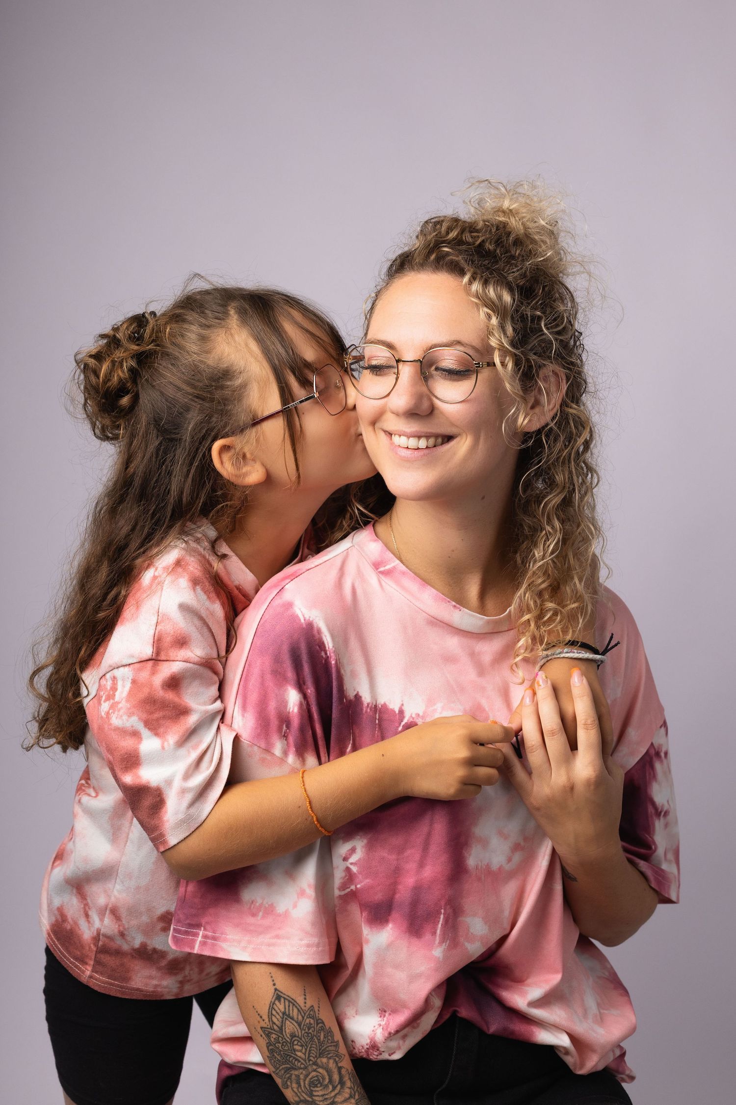 Young girl kissing smiling curly-haired woman wearing matching pink tie-dye shirts in studio photo.