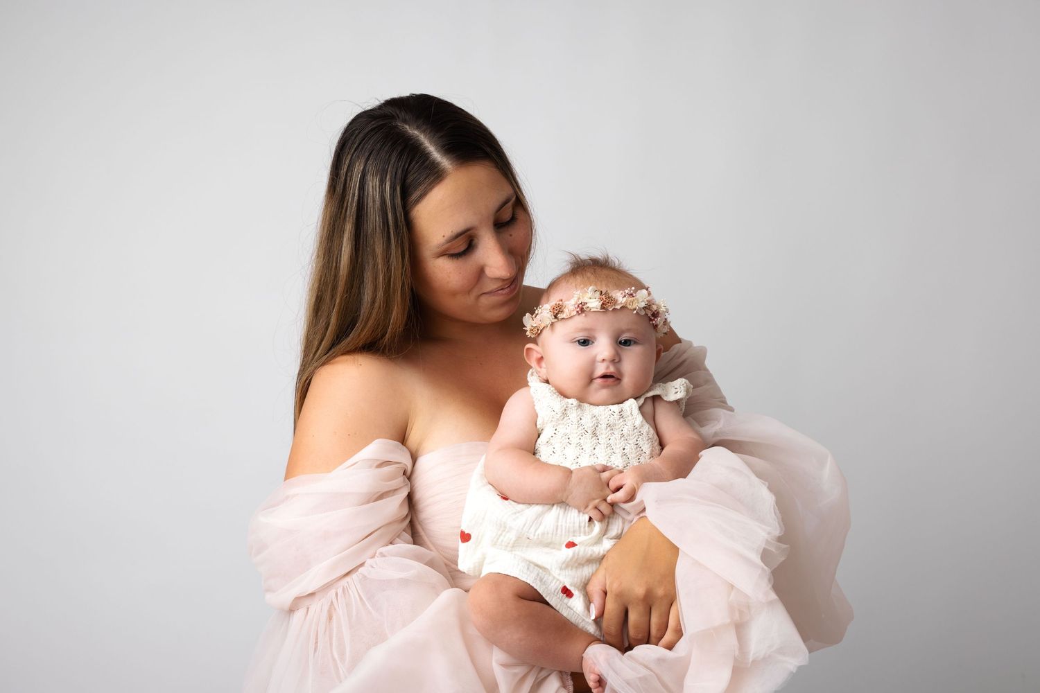 A mother in a pink gown tenderly holds her baby girl in a white dress and floral headband against a white backdrop.