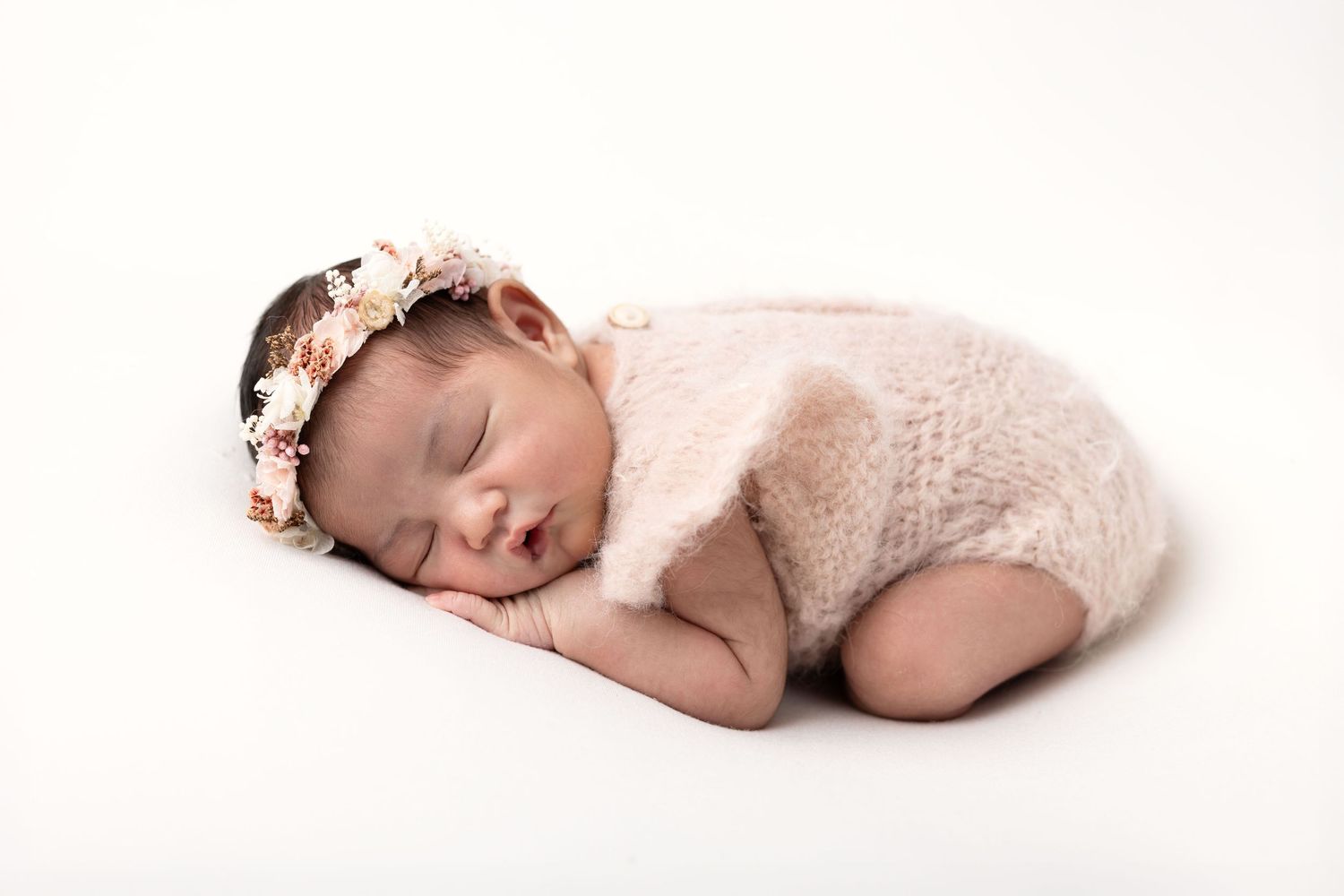 Sleeping newborn baby girl in pink knit romper and floral headband on white background.