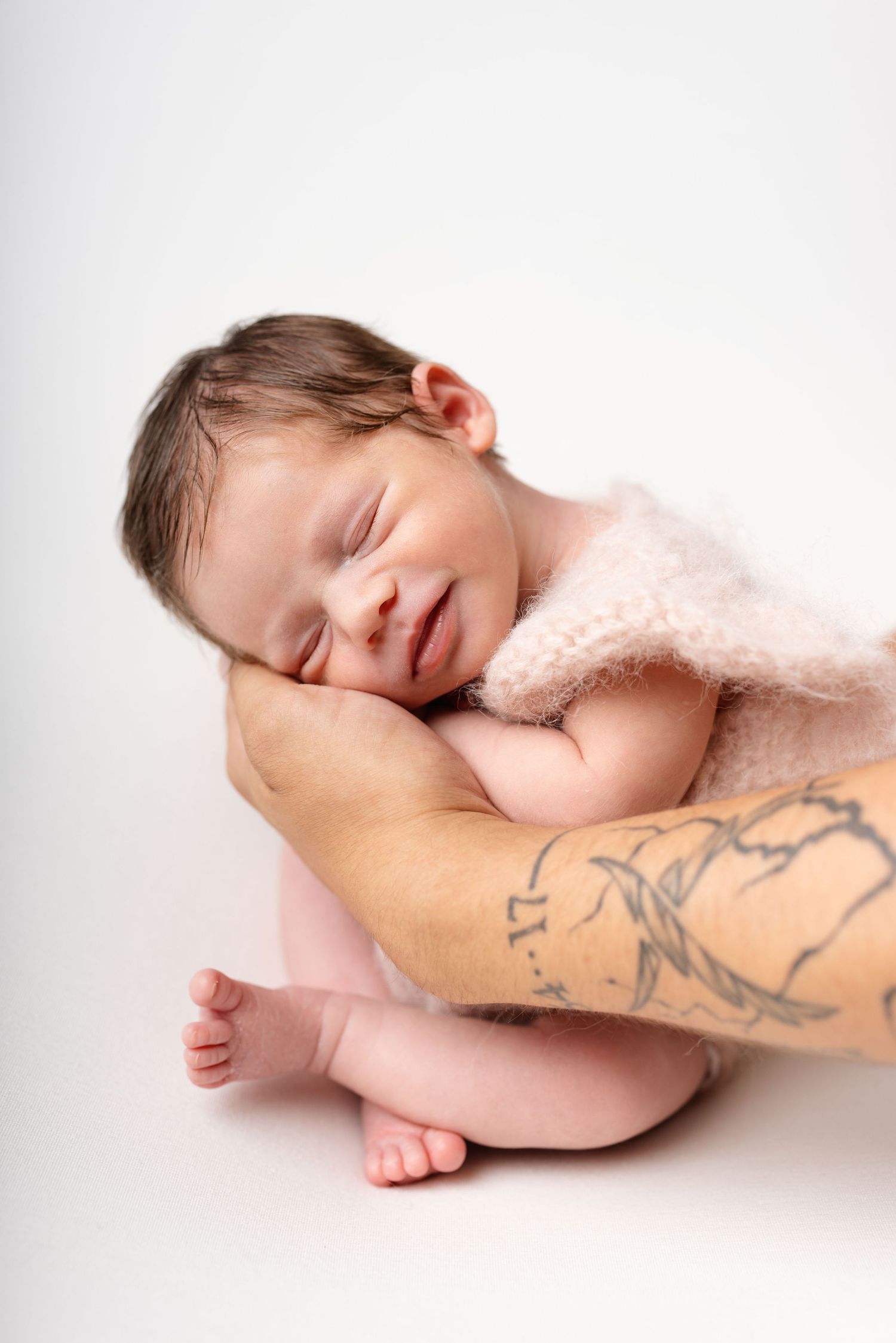 Newborn baby sleeping on tattooed arm, wrapped in pink blanket, smiling peacefully against white background.