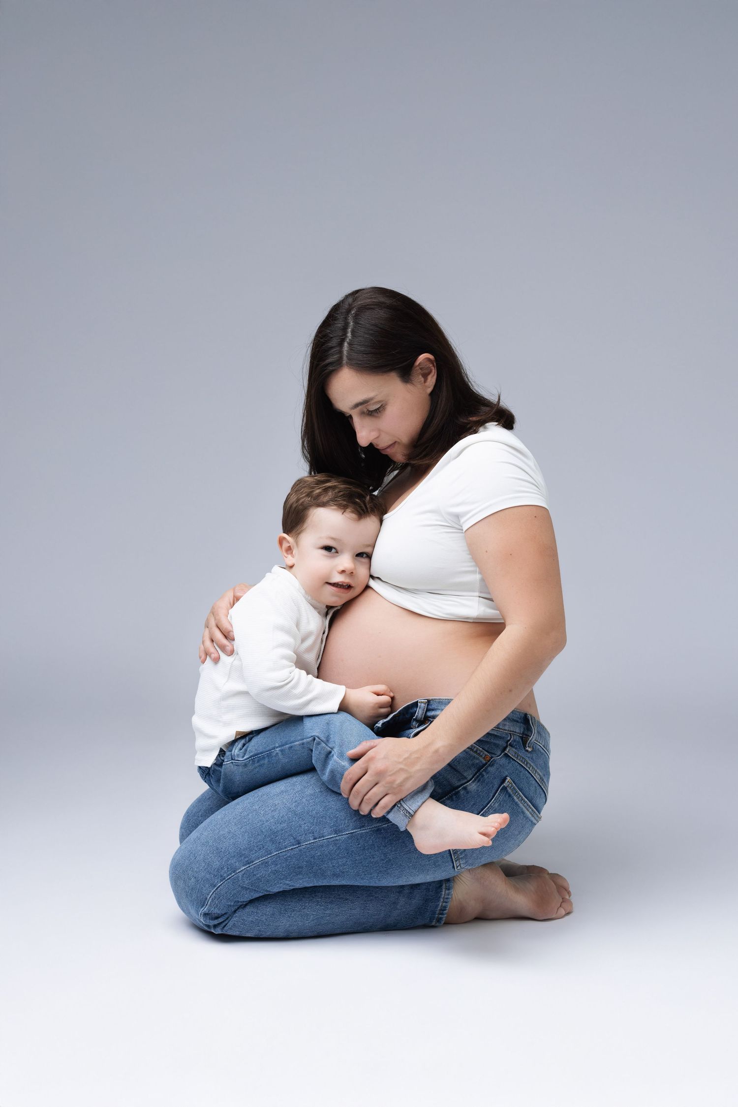 Pregnant woman in white top and jeans sitting with toddler son hugging her baby bump. photographe grossesse chavanoz lyon