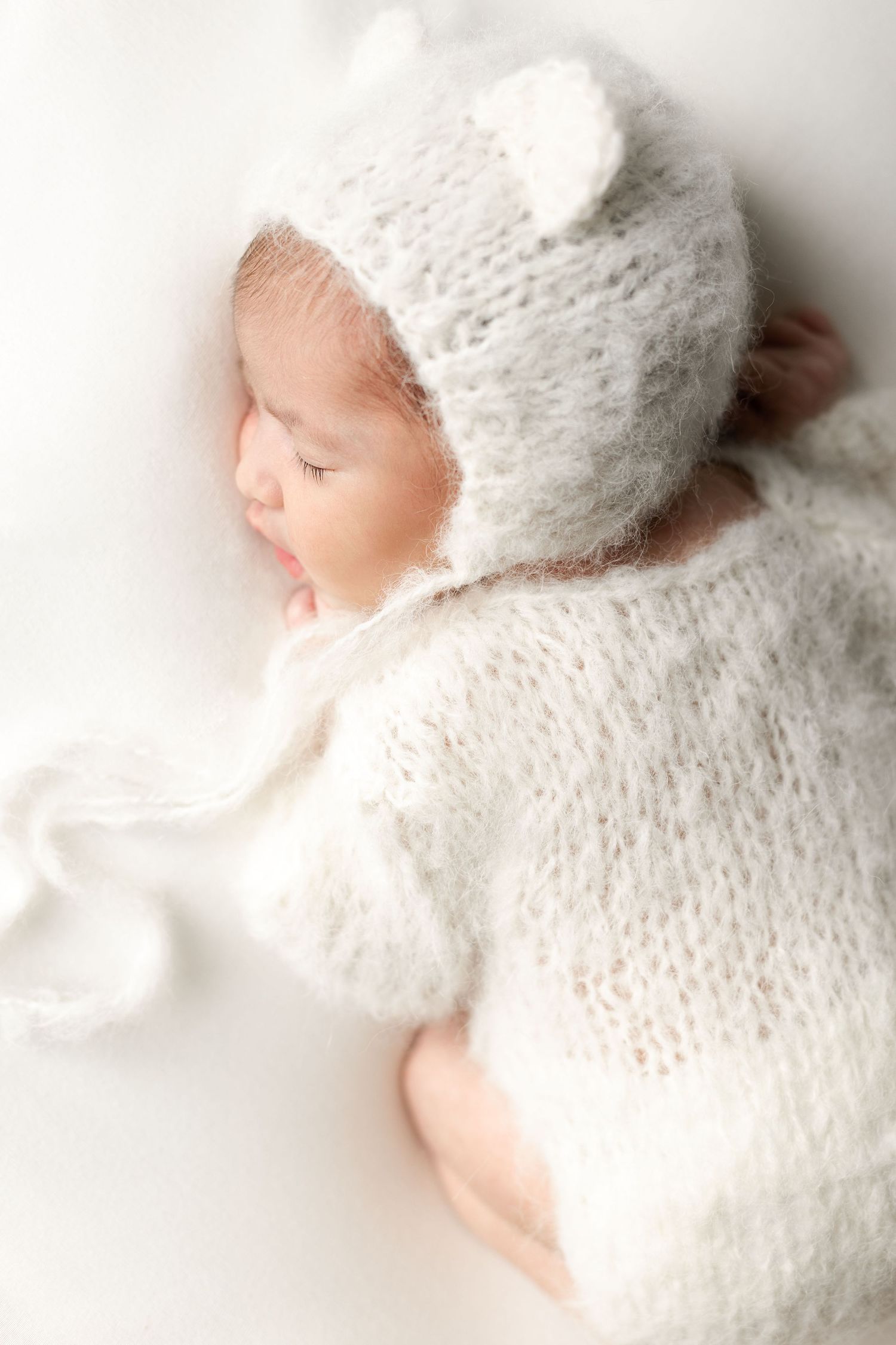 Close-up of a sleeping newborn wearing a soft white mohair knit romper and matching bear ear bonnet. séance naissance chavanoz lyon