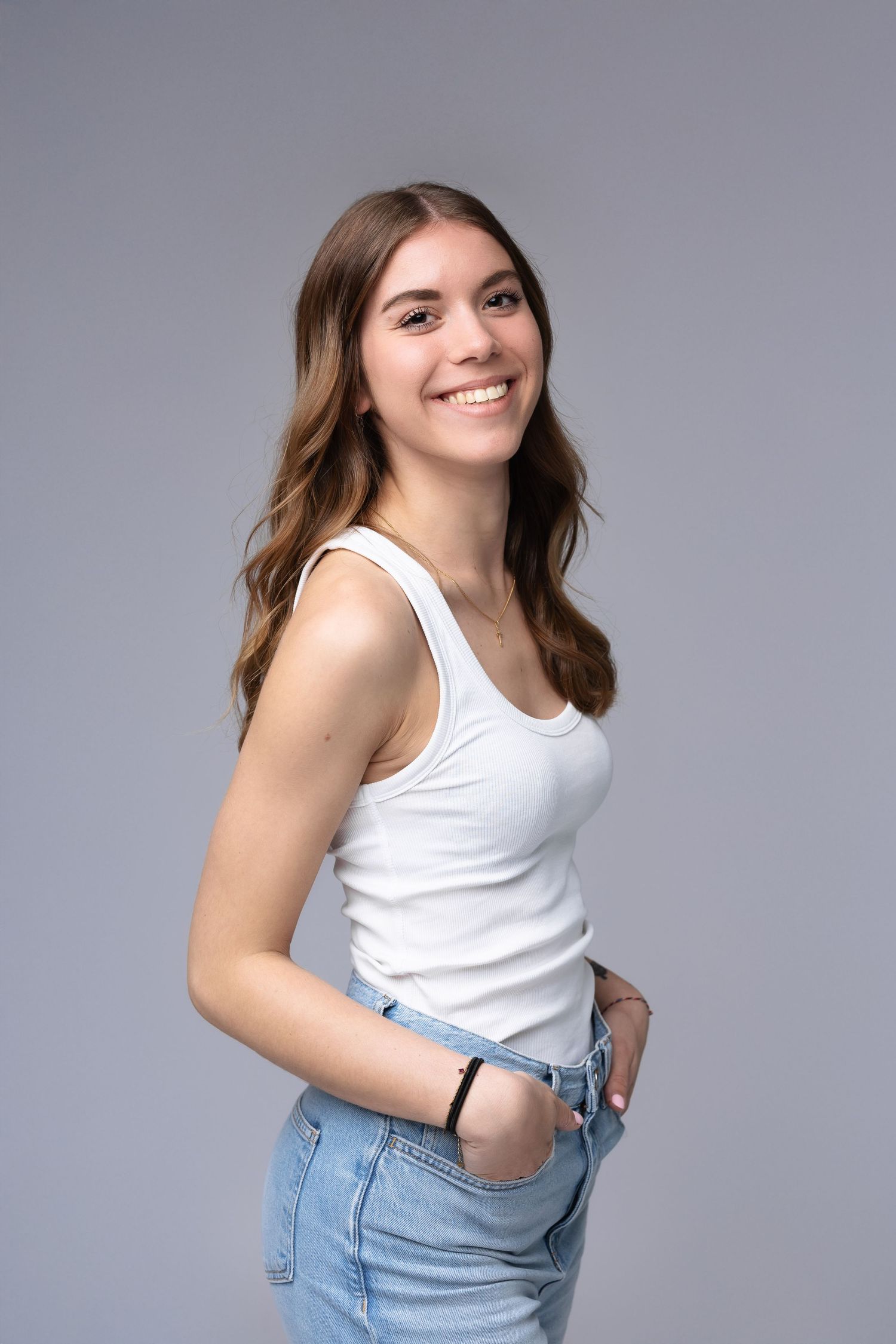Smiling young woman in white tank top and jeans posing confidently in studio portrait.