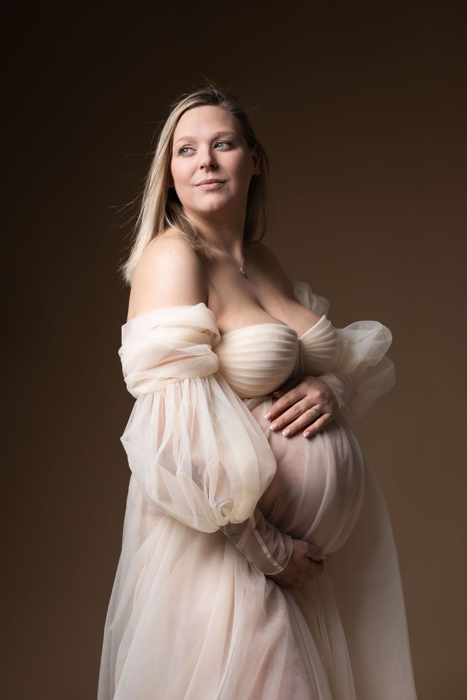 Smiling blonde pregnant woman in sheer cream tulle gown cradles bump in warm studio light.