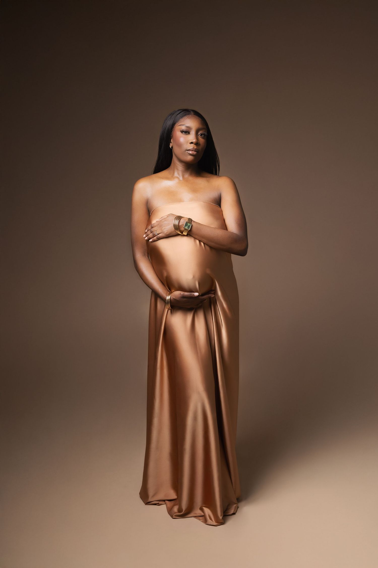 Pregnant Black woman draped in gold satin fabric posing in studio.