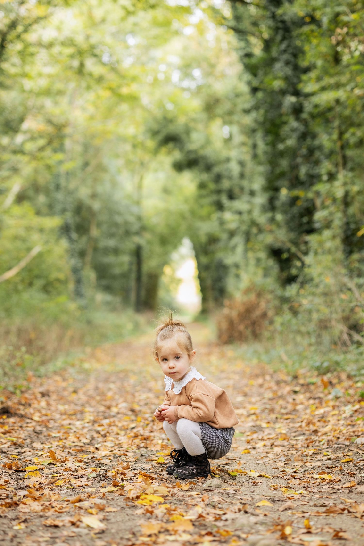 Toddler in brown jacket crouching on a leaf-covered path in an autumn forest tunnel.