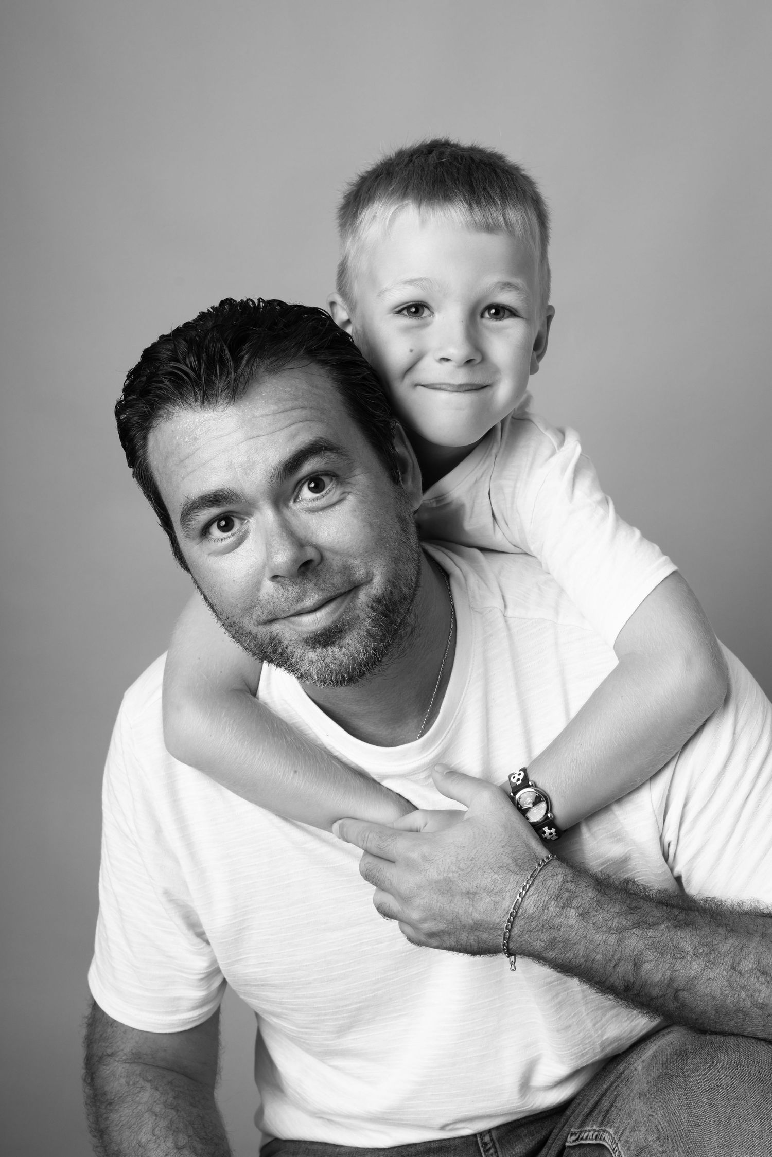 Black and white portrait of a smiling man and young boy hugging, wearing white t-shirts against a neutral background.