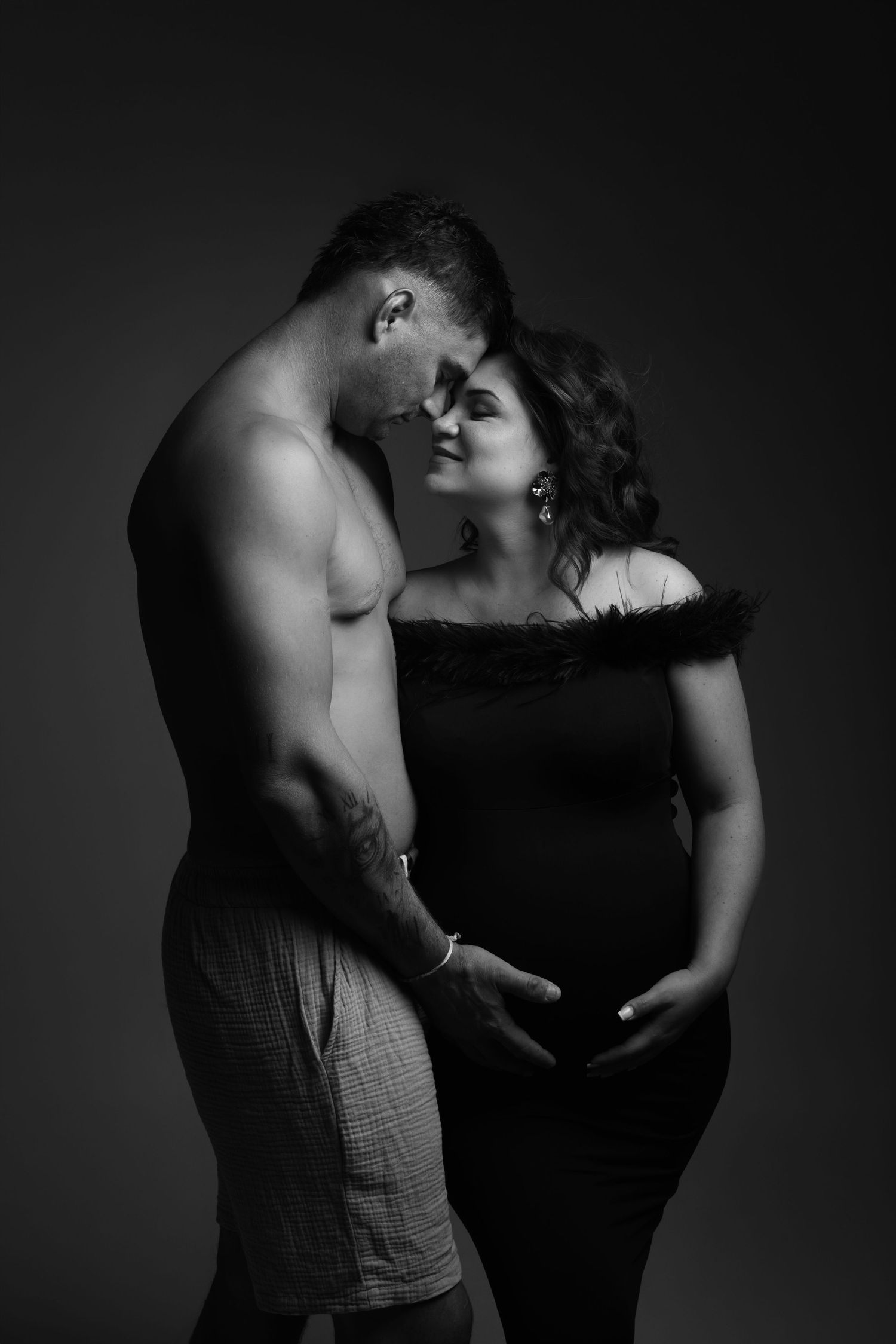 Black and white photo of a shirtless man tenderly embracing a pregnant woman in a studio maternity portrait.